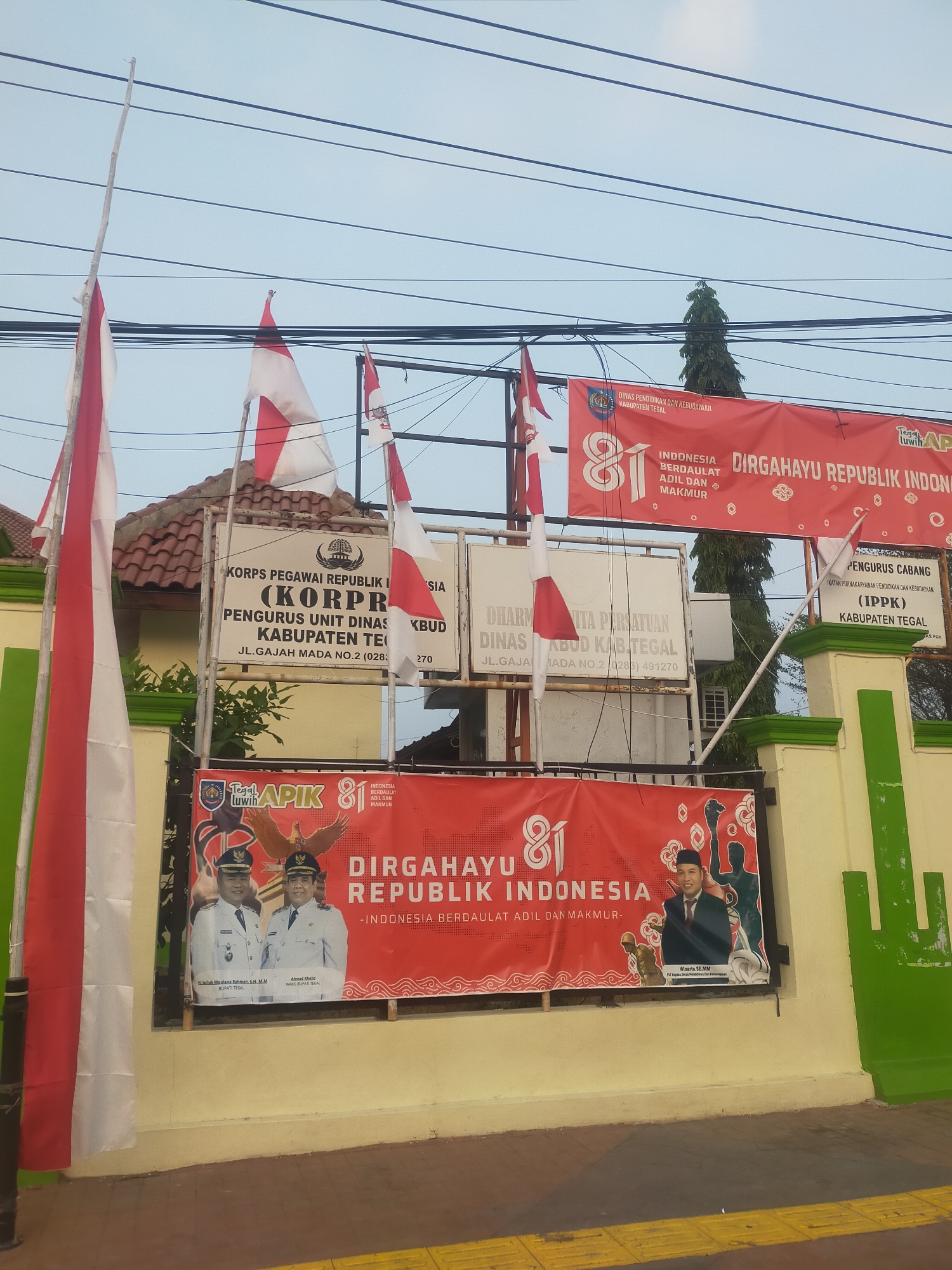 The image depicts the exterior of a building in Slawi, Indonesia, likely a government office or educational institution, adorned with patriotic decorations. Two large red and white Indonesian flags are prominently displayed on the left, one on a tall pole and another attached to the building. Multiple banners are hung across the facade.
The main banner, stretching across the front, is red with white lettering that reads "DIRGAHAYU REPUBLIK INDONESIA" (Long Live the Republic of Indonesia) and "81", signifying the 81st anniversary of Indonesian independence. Smaller banners on the left display the emblems of the "KORPS PEGAWAI REPUBLIK INDONESIA (KORPRI)" and "DINAS PENDIDIKAN DAN KEBUDAYAAN KABUPATEN TEGAL" (Education and Culture Office of Tegal Regency). Another banner on the right refers to "PENGURUS CABANG IKATAN PURN... KEPENDIDIKAN DAN KEBUDAYAAN (IPPK)". Below the main banner, a smaller red banner features portraits of two men in uniforms, identified as "BUPATI TEGAL" (Regent of Tegal) H. UCHAK MAULANA RAHMAN, S.H., M.M. and AHMAD CHOIR, S.H., M.H. (implied to be another official). To the right of these portraits is another portrait of an individual in a suit and tie, identified as WISARTO, S.E., M.M. R/KT KEPALA DINAS PENDIDIKAN DAN KEB..." (likely a head of the Education and Culture Office).
The building's facade is painted in shades of yellow and light green. Wires run overhead, and a tall, dark green tree is visible behind the banners. The ground in the foreground appears to be a sidewalk with yellow tactile paving, indicating accessibility features. The overall scene suggests a festive atmosphere celebrating Indonesian Independence Day.