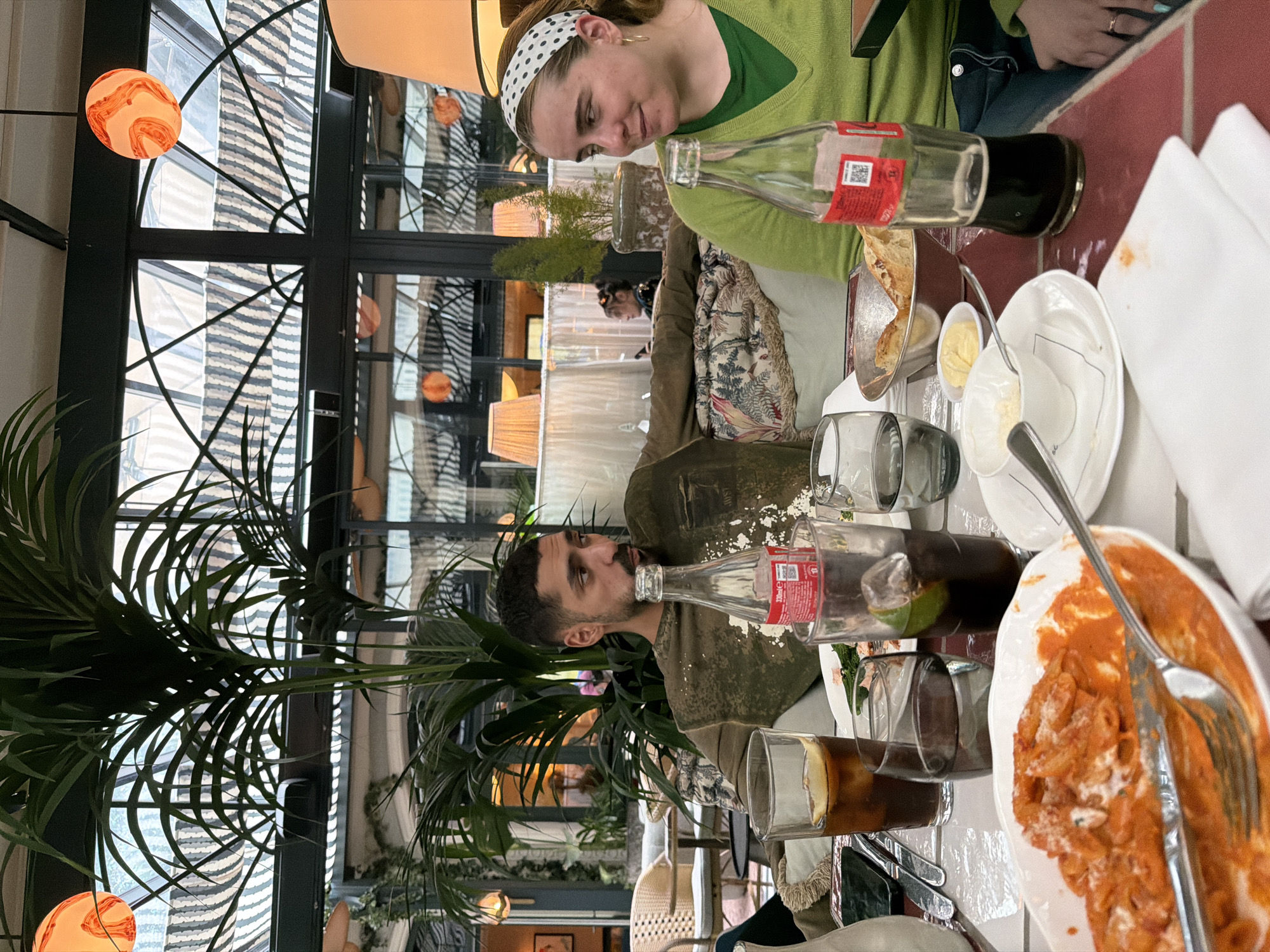 A photograph depicts two people seated at a table in a restaurant, likely in Paris, France, based on the style of the architecture and décor. A woman with a white and black spotted headband wears a green sweater. A man is wearing a green shirt. The table is set with plates of pasta, glasses of drinks, and Coca-Cola bottles. A large leafy plant obscures a portion of the view behind them. The restaurant features large windows with curved metal detailing and hanging spherical lights.