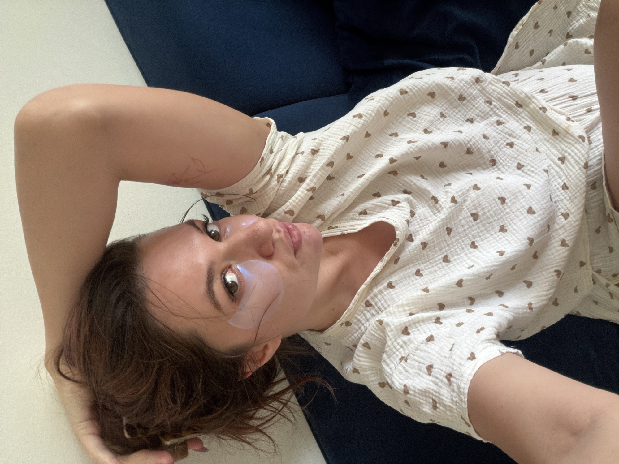 A woman lies on a blue sofa, facing the camera. She is wearing a light-colored dress with small brown hearts printed on it and has hydrogel eye patches on. Her hair is brown. The location is unknown and there is nothing in the image to suggest it is Belgrade, Serbia.