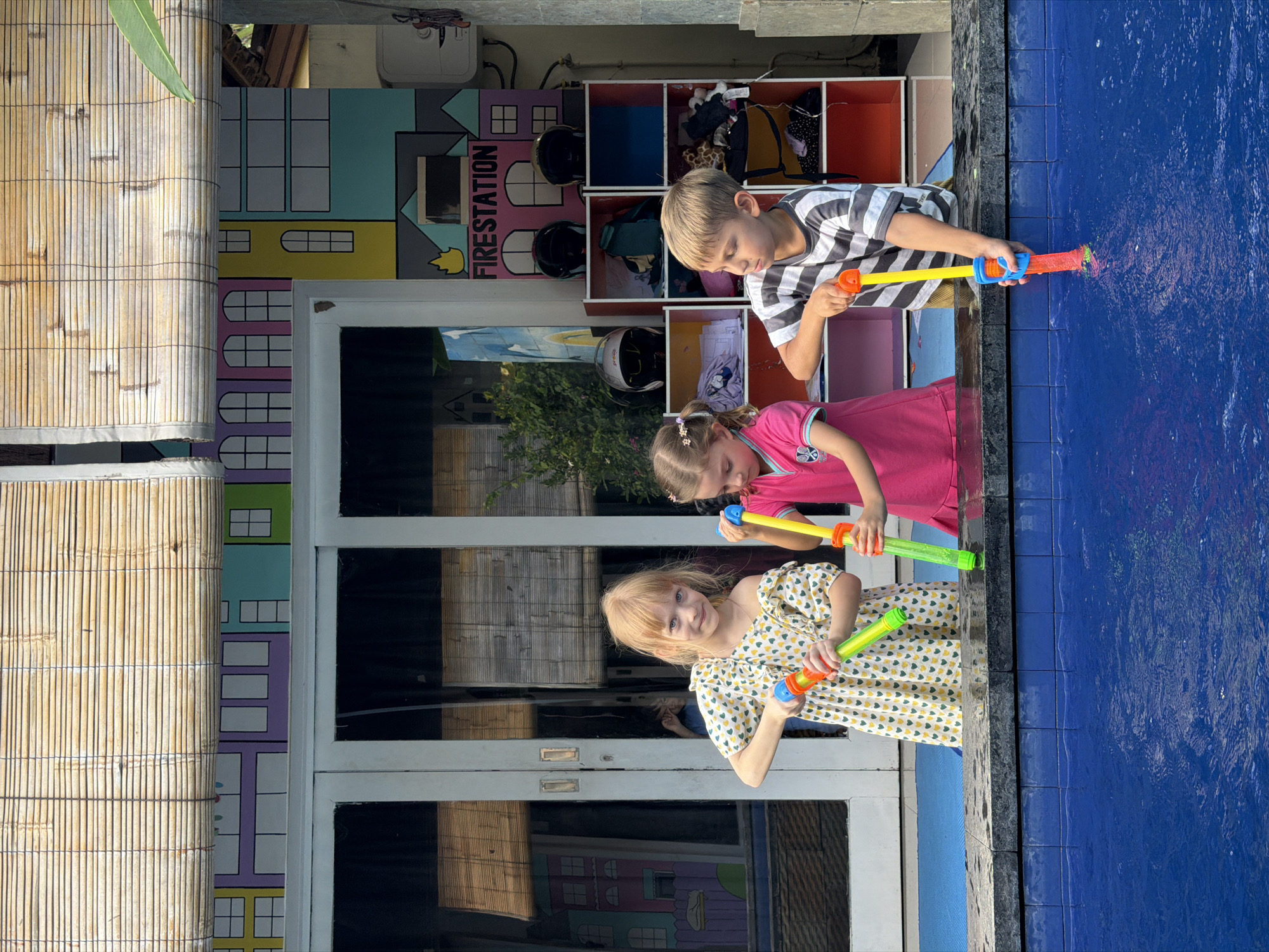 Three children are playing with water guns alongside a pool. A boy in a striped shirt and two girls, one in a pink shirt and one in a patterned dress, are aiming the water guns at the pool. Behind them, there's a building with colorful, cartoon-like paintings of buildings and a "fire station" sign. There is also a cabinet storing motorcycle helmets, clothes, and other items. There is a bamboo wall on the left side. The location is likely in Kuta, Indonesia, based on the architectural style and the tropical environment.
