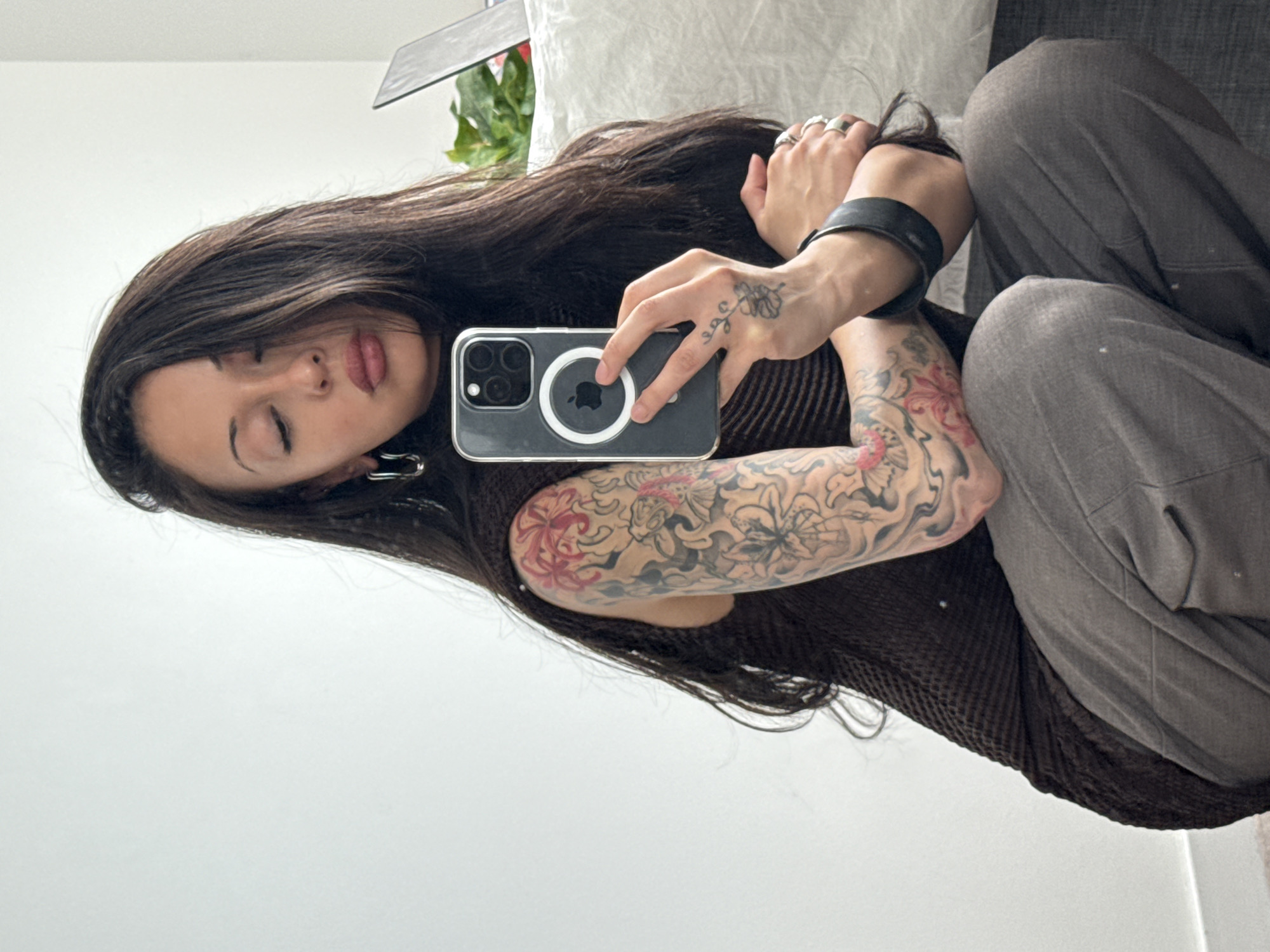 The image captures a person taking a mirror selfie indoors, likely in a private residence in London, United Kingdom. The subject, who appears to be female, is seen from the chest up, with long, dark hair flowing over her shoulders. Her head is tilted, and her eyes are closed, conveying a sense of calm or introspection.
She is wearing a dark brown, possibly ribbed, sleeveless top and light grey trousers. Her left arm is heavily adorned with intricate tattoos featuring floral and abstract designs in shades of red, grey, and black. A smaller, delicate tattoo resembling a plant or floral motif is visible on the back of her right hand. On her left hand, she wears multiple rings, and a thick, black wristband is on her left wrist.
The person holds a grey smartphone, identifiable as an iPhone due to the prominent white Apple logo and MagSafe circle on its back, in her right hand, pointing towards herself to capture the reflection. The background includes a plain light-colored wall, a glimpse of a grey sofa or upholstered furniture, and a green houseplant, indicating a relaxed indoor setting. The lighting is soft and even, suggesting an interior during the daytime or a well-lit evening. No other visible text is present.