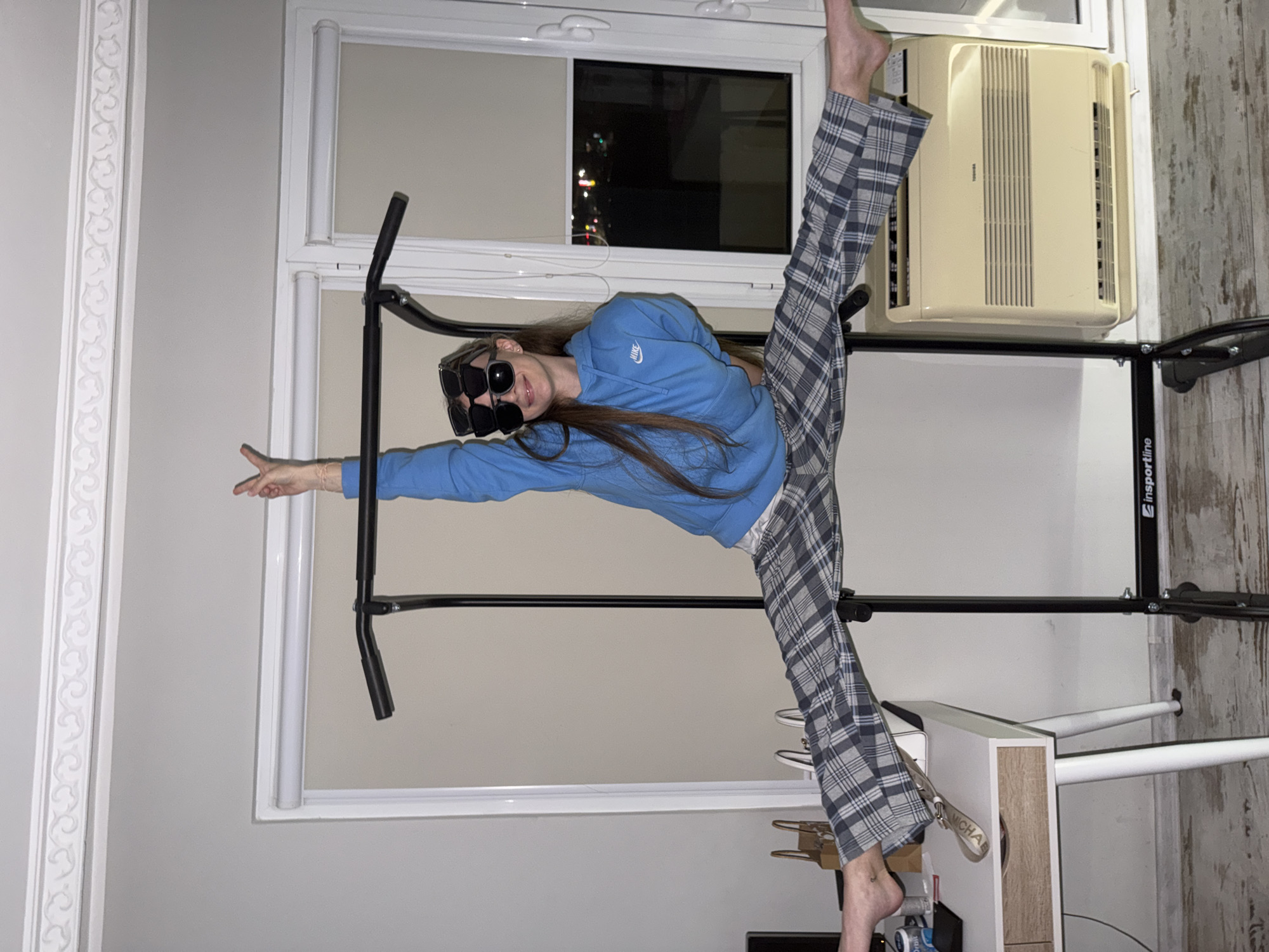 A rotated, eye-level indoor shot shows a female with long brown hair in a blue Nike hoodie and grey plaid pants doing a split between a window with a black metal pull-up bar and a white desk. She is wearing multiple pairs of black sunglasses stacked on her face. Her right arm is extended to the left, and her left leg is extended up. Her right leg is angled downward. An air conditioning unit is situated above the pull-up bar. The brand “Sportline” is visible on the pull-up bar. A key chain with the name “MICHAEL” is attached to the desk. The location appears to be inside of a home. The lighting is bright. The location in the city of Plovdiv, Bulgaria is not determined in this image.