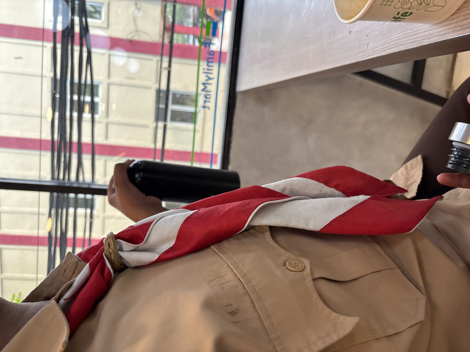 A person wearing a tan scout uniform with a red and white scarf around their neck is holding a black water bottle. The person is seated at a wooden table, and in the background, there is a window with a view of a building. The building's facade features vertical pink stripes. A sign on the window reads "FamilyMart". There is a paper cup on the table to the right of the person. The scene is indoors, possibly in a cafe or a similar establishment in Binong, Indonesia.