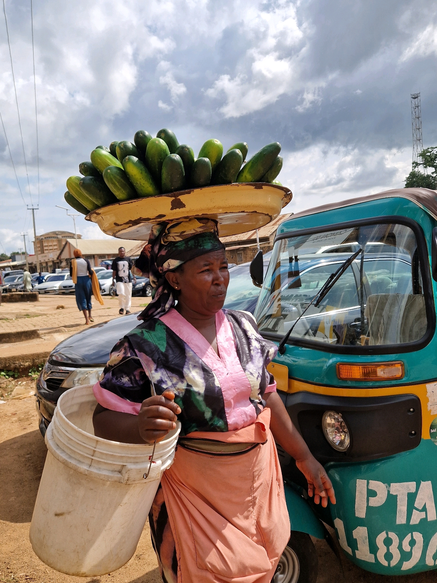 An outdoor, medium shot, eye-level, vertical photograph captures a woman carrying a large load of cucumbers on her head in Jos, Nigeria. The woman, who is Black, is wearing a colorful headscarf and a pink and dark patterned dress. She is holding a white bucket in her right hand. She is standing in front of a teal and yellow three-wheeled vehicle, which is common in Nigeria and is called an "okada". In the background, other vehicles are parked, and buildings are visible, suggesting a market or roadside scene. The sky is cloudy. The woman's expression is neutral. The overall lighting suggests it is daytime.