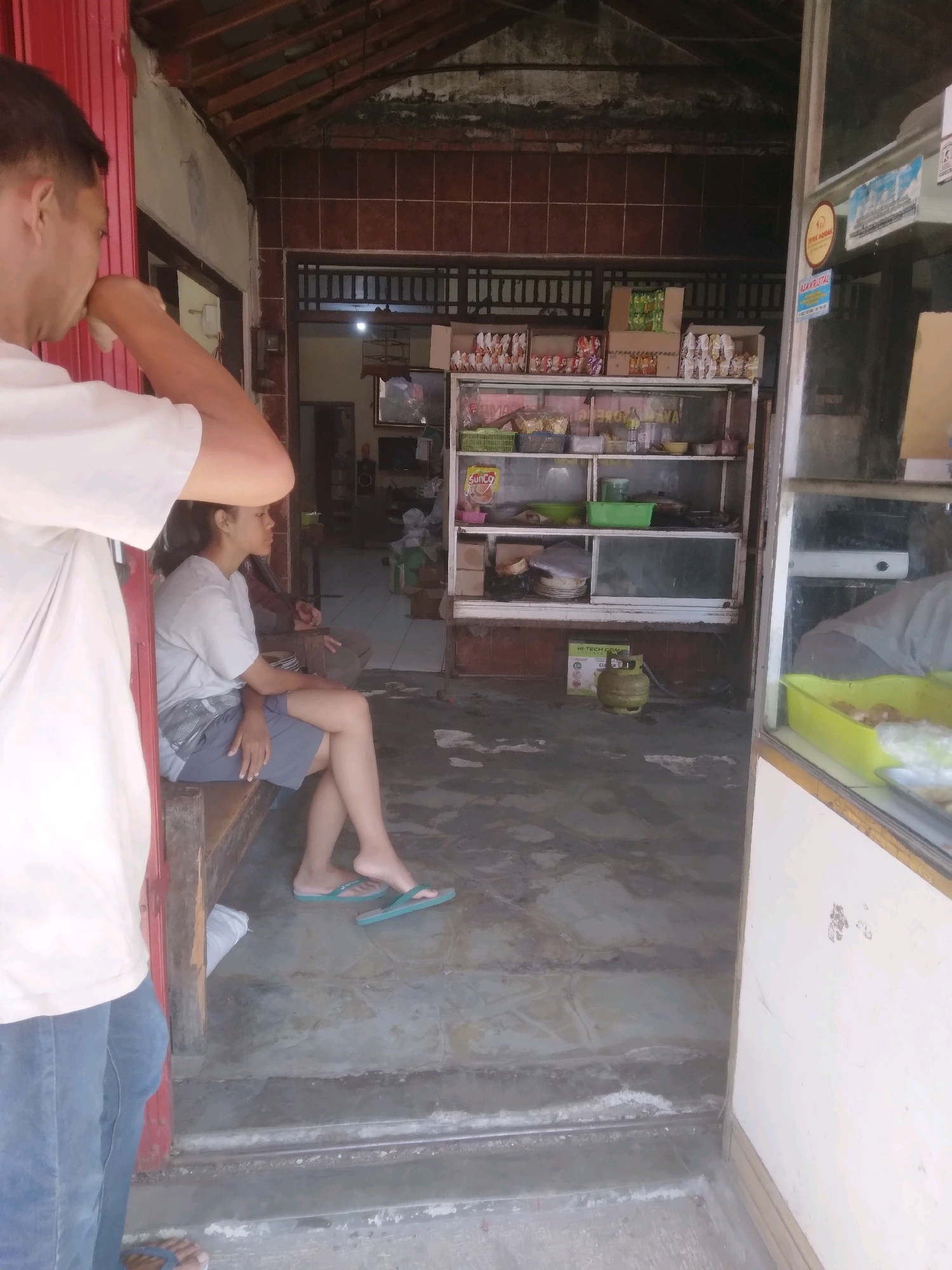 The image depicts the interior of a small shop or eatery in Purwokerto, Indonesia. A person, partially visible on the left, is wearing a white t-shirt and blue jeans. In the center, a woman is seated on a wooden bench, wearing a grey t-shirt and dark shorts, with her legs crossed and wearing flip-flops. She is looking to the right. Behind her, another person is seated on the same bench, but their face is not visible. The environment appears to be an open-air establishment with a tiled floor and a wooden ceiling. Shelves are visible inside the shop, stocked with various food items and containers. A yellow gas cylinder is placed on the floor near the shelves. The scene suggests a casual, possibly informal business setting.