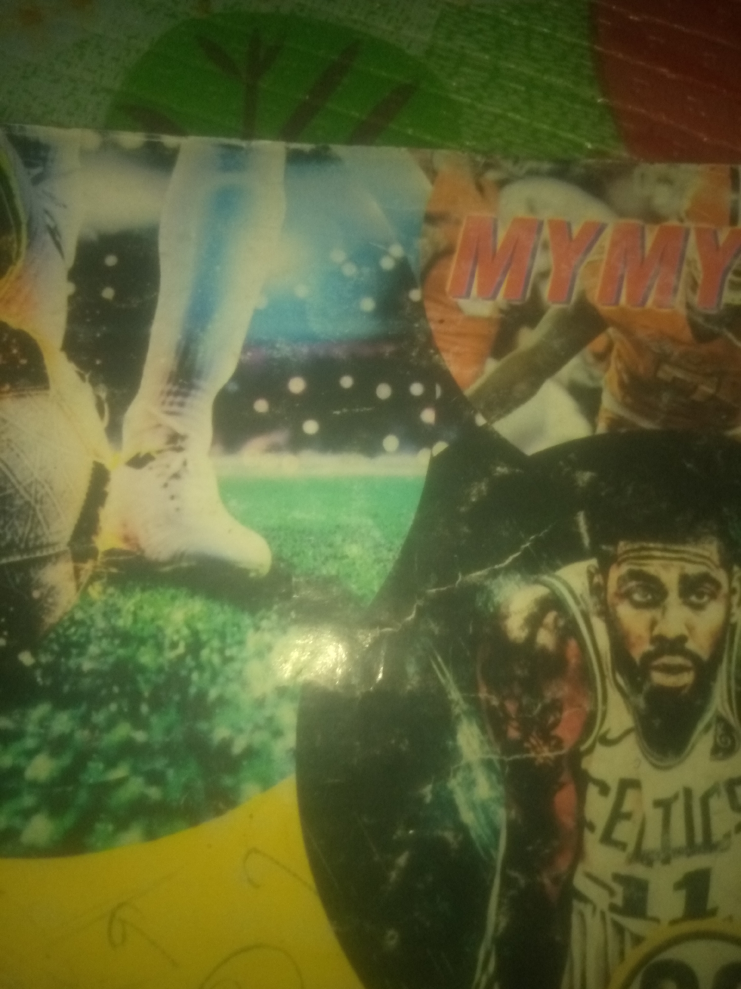 The image is a close-up of a poster or magazine cover featuring basketball players. In the foreground, on the right side, a close-up shot of a basketball player's face and torso is visible. He is wearing a white jersey with green and yellow trim and the word "CELTICS" is emblazoned across his chest, along with the number "11". He has dark skin, a beard, and is looking towards the viewer. To the left of this player, another basketball player's legs are visible, one leg is extended forward with a basketball at his feet. In the upper portion of the image, the word "MYMY" is printed in large orange letters. The background consists of abstract colorful shapes, possibly representing a stadium or crowd. The image appears to be aged and somewhat damaged, with visible creases and scuff marks. There is no discernible information or visual cues within the image to suggest a location in Jauro Yinu, Nigeria.