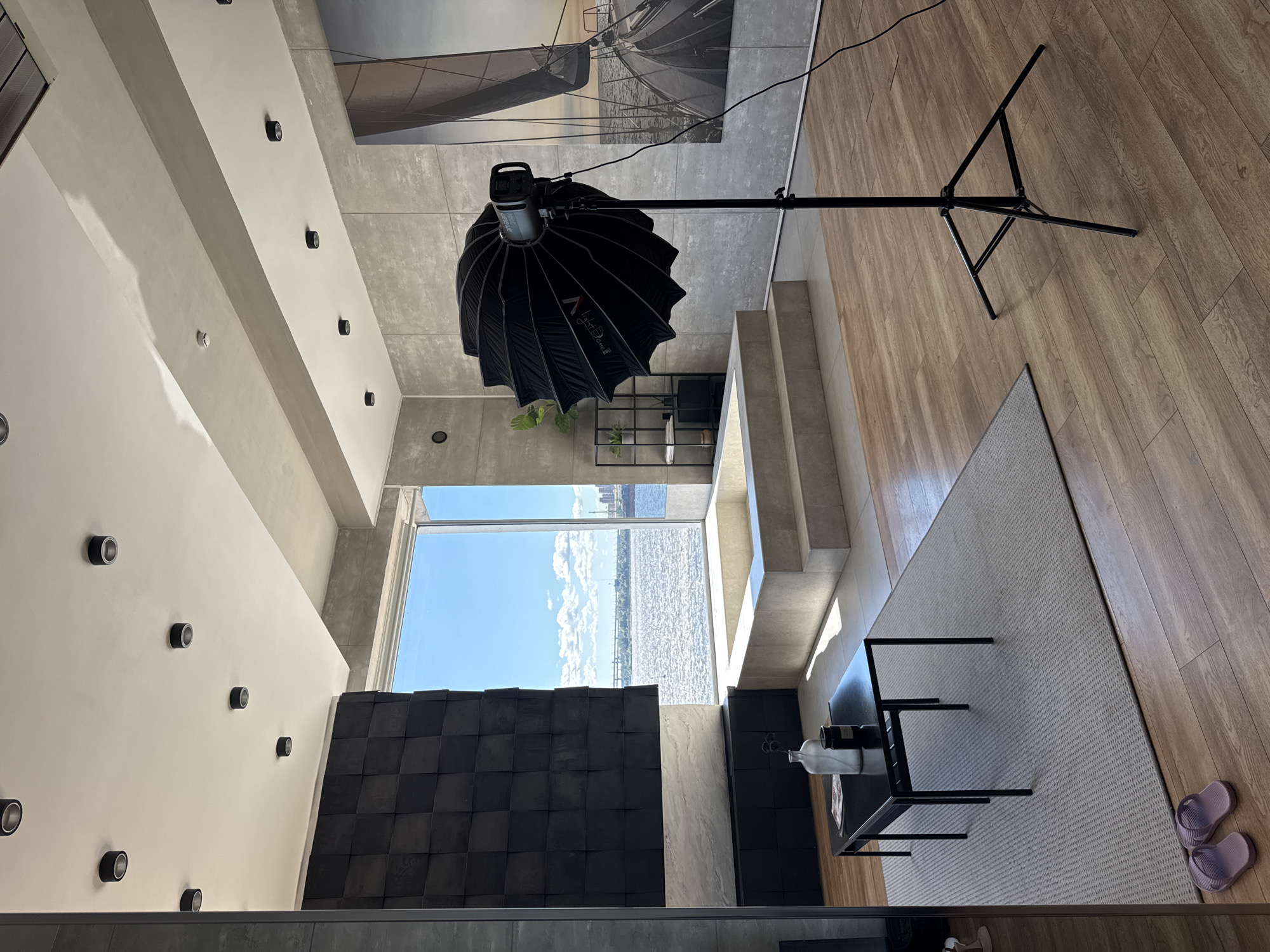 This image depicts an interior scene of a modern apartment with a large window overlooking a body of water, likely the Neva River in Saint Petersburg, Russia, given the architectural style and waterfront view. The room features a raised bathtub area with tiled steps, a black coffee table on a grey rug with two bottles and a candle, and a pair of purple flip-flops on the floor. A large black umbrella-like light modifier on a stand is positioned in the foreground to the right, suggesting a photoshoot is taking place. The walls are decorated with large grey tiles, and a textured black wall panel is visible to the left. A large artwork of a sailboat is mounted on the wall on the right. The ceiling is adorned with multiple recessed lights. The overall atmosphere is contemporary and minimalist, with a focus on natural light and the expansive water view. There are no people present in the image.