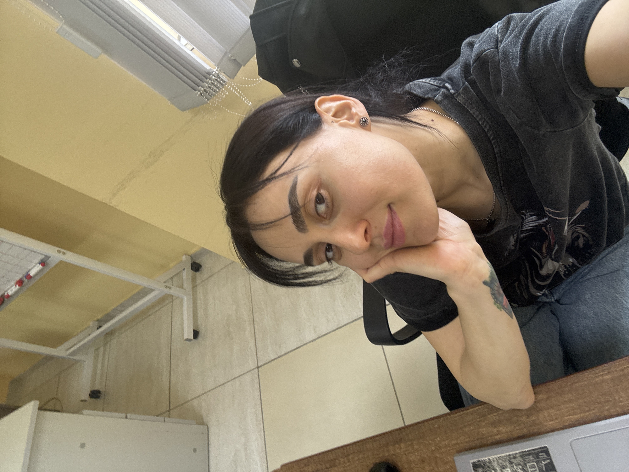 A medium, angled indoor shot depicts a woman resting her head on her hand. She has dark hair, fair skin, and wears a dark-colored shirt, jeans, and a silver chain necklace. There is a small tattoo visible on her arm. A black backpack is behind her.
The environment consists of a room with tiled floors and yellow walls. A white desk with a laptop on it is in the foreground. Window blinds are visible at the top of the frame.
The location is unknown.
