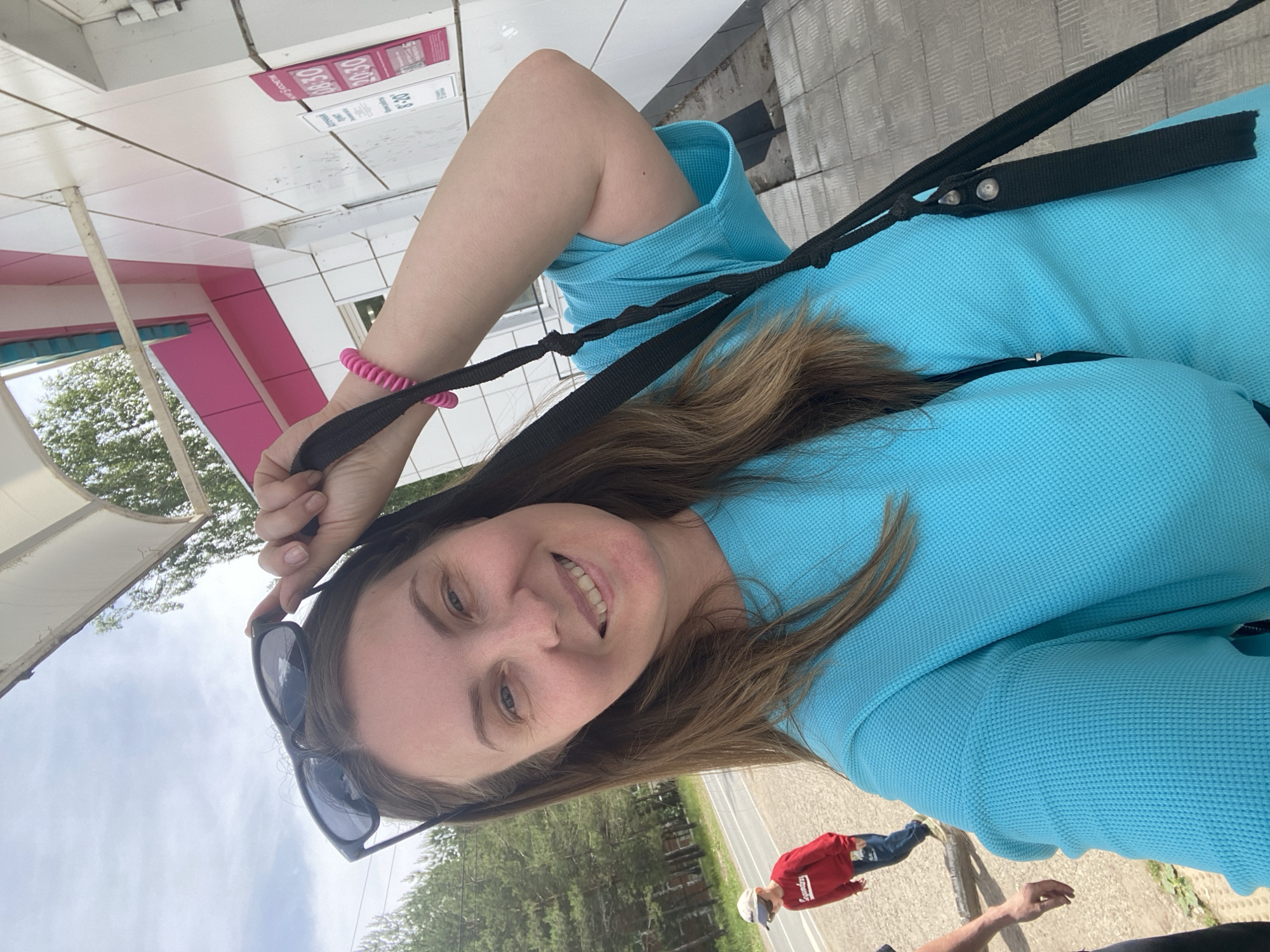 A woman is featured in a brightly lit, outdoor selfie, rotated 90 degrees clockwise from a typical upright orientation. She smiles at the camera, displaying light brown hair, blue eyes, and fair skin. She is wearing a textured, light blue long-sleeved top and has a pink coil bracelet on her left wrist. Black sunglasses rest on her head, and she holds a black strap, possibly from a bag or purse.
The background reveals a building with a distinctive pink and white facade, likely a shop or kiosk. A sign on the building displays visible white text including "08:30", "20:30", and "8:20", which appear to be time indicators such as opening hours. Beyond the building, a paved road is visible, flanked by green trees. Another individual, possibly a child, wearing a red top and blue pants, stands in the mid-ground. Part of a third person's arm and hand can be seen in the lower right corner of the frame.
The scene depicts a casual daytime moment in Noviy, Russia, with clear lighting suggesting sunny or partly cloudy weather. The woman's expression conveys happiness and ease.