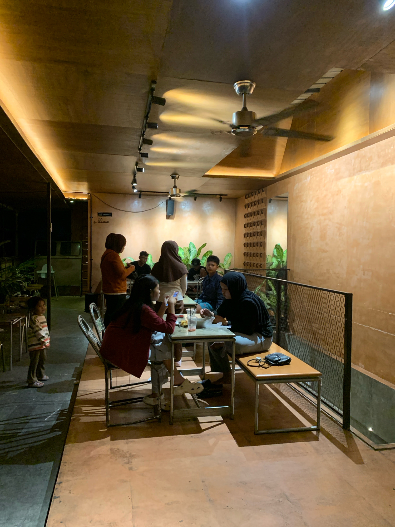The image depicts a dimly lit, open-air cafe with a rustic ambiance, likely located in Mojokerto, Indonesia, given the context. The ceiling features exposed wooden beams with track lighting and a ceiling fan, contributing to the industrial-chic aesthetic. The walls are textured and painted in a warm, earthy orange hue, with a patterned decorative element on one wall.
Several individuals are present, seated at tables and interacting. In the foreground, two young women are seated at a table, one looking at her phone, the other with her back to the camera, engaged in conversation or eating. In the mid-ground, a group of people are seated at another table, including men and women, some appearing to be engaged in conversation. A child in a striped shirt stands to the left of the frame, looking towards the cafe entrance.
Objects in the scene include tables with drinks and plates, chairs, and a bench with a bag on it. The flooring is a mix of concrete and what appears to be wood or a similar material, with strong shadows cast by the lighting. Lush green plants are visible in the background, adding a touch of nature to the urban setting. The overall atmosphere suggests a casual gathering place, possibly a popular spot for locals to socialize.