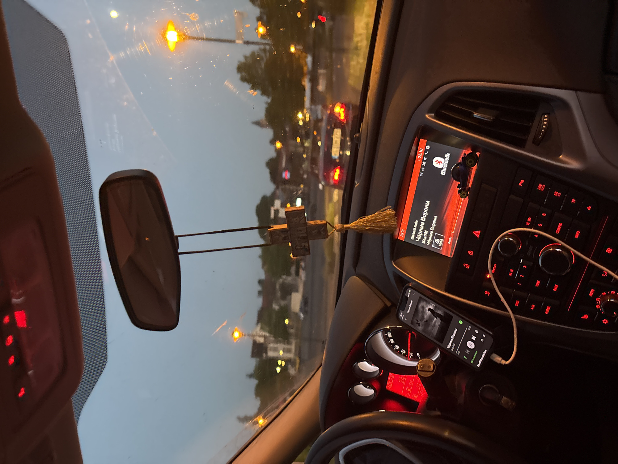 The image shows the interior of a car at dusk, with the view through the windshield. The dashboard is illuminated with red and orange lights. A smartphone is mounted on the dashboard and displays a music player interface. A rosary hangs from the rearview mirror. Outside the car, streetlights and building lights are visible. Another car is driving ahead. The overall impression is that of a car journey at twilight.