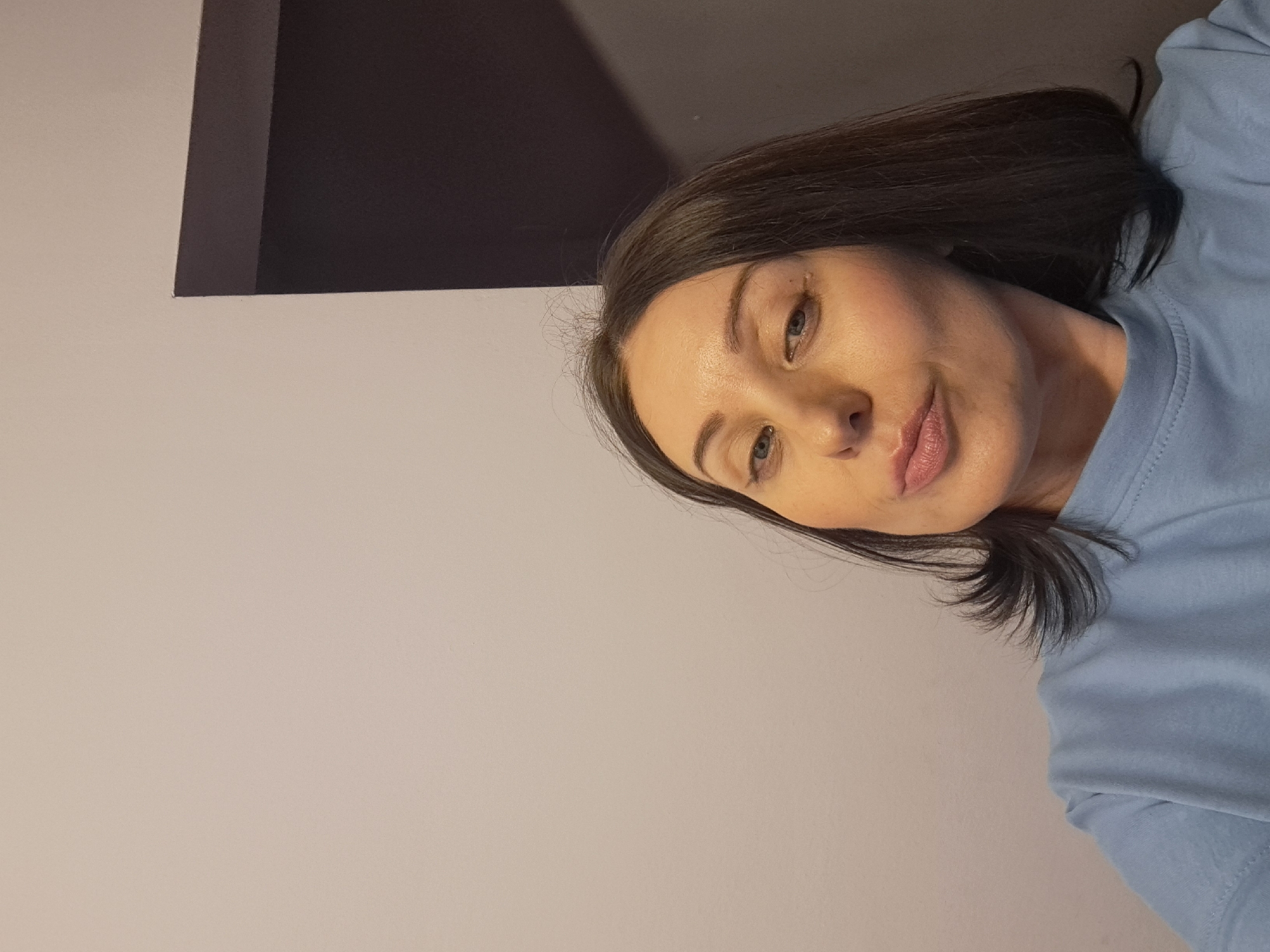 A woman is pictured from the chest up, with her head turned to her right. She has brown hair, blue eyes, and is wearing a light blue t-shirt. The background is a plain, light-colored wall with a darker rectangular shape on the right side of the image, possibly a doorway or a piece of furniture. The lighting appears to be artificial and coming from the front. The woman is making a pouty expression. The image is a selfie, likely taken indoors. There is no discernible information in the image to suggest a specific location in Irkutsk, Russia.
