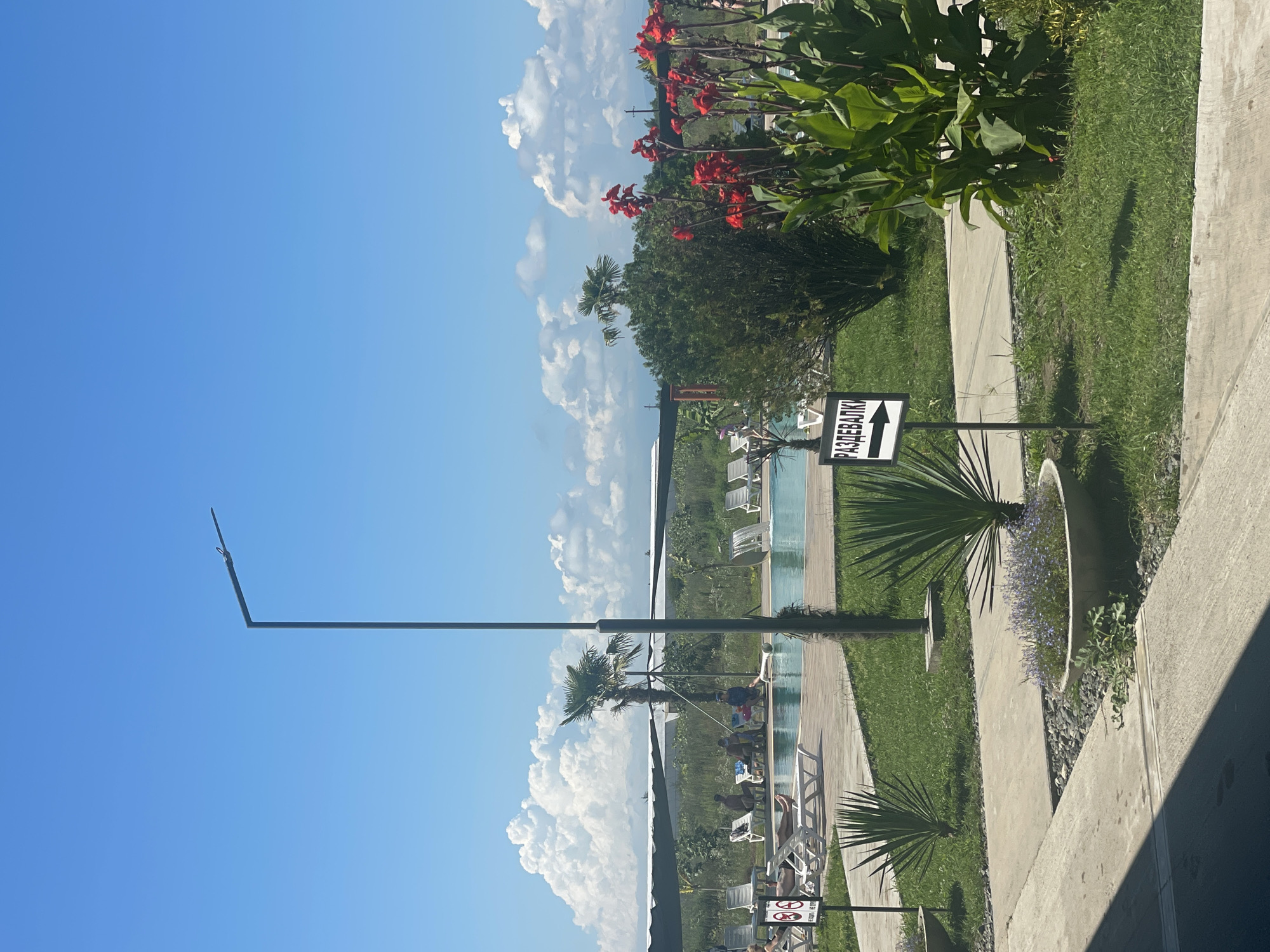 The image shows a sunny day scene at an outdoor pool area. A tall, modern lamppost stands prominently in the foreground, casting a shadow. To the right, vibrant red flowers bloom among lush green foliage. A sign with an arrow pointing to the right indicates "РАЗДЕВАЛКИ" (Changing Rooms). In the middle ground, a swimming pool is visible, with several people relaxing on lounge chairs by the poolside. Palm trees and other greenery dot the landscape, with a backdrop of white, puffy clouds against a clear blue sky. The scene appears to be in a resort or recreational area in Adzyubzha, Abkhazia.