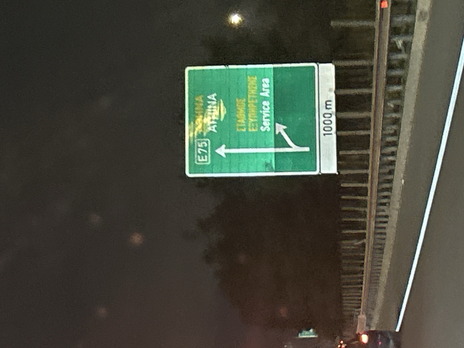 The image captures a highway sign at night, oriented vertically due to the original rotation. The primary subject is a large, reflective green overhead sign, characteristic of European motorways. To its right, a narrower white sign provides additional information. The scene is dark, indicating nighttime, with faint glints of light suggesting reflections from streetlights or passing vehicles, and possibly some lens flare.
The green sign displays directional information in both Greek and English. On the left side, an arrow pointing straight ahead or slightly left indicates "E75" and "ΑΘΗΝΑ ATHINA" (Athens). On the right side, a diverging arrow points towards "ΣΤΑΘΜΟΣ ΕΞΥΠΗΡΕΤΗΣΗΣ Service Area." This suggests a fork in the road where drivers can continue on European Route E75 towards Athens or take an exit for a service area.
The adjacent white sign clearly displays "1000 m," indicating that the upcoming junction or service area is 1000 meters away. A metal guardrail or barrier runs along the right side of the frame, separating the road from the surrounding area. The road surface below the sign is dimly visible. The overall impression is of a driver's perspective approaching a highway exit or service area at night in Agios Stefanos, Greece. No people are visible.