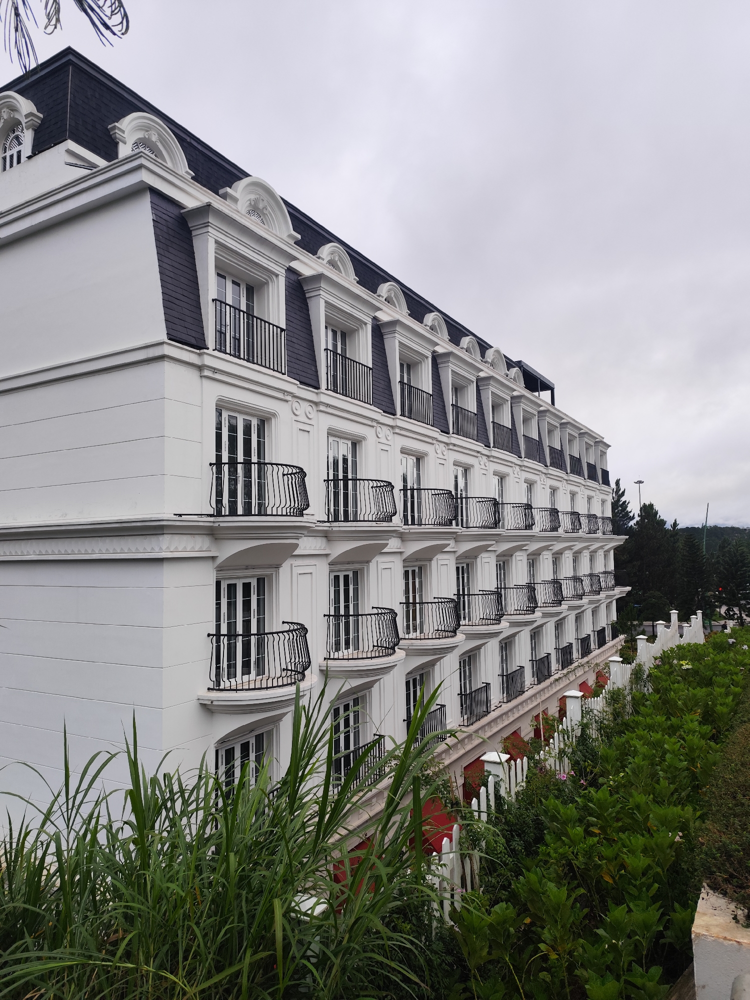 The image shows a multi-story white building with a dark mansard roof, featuring multiple windows and small balconies with black wrought-iron railings on each level. The architecture suggests a European influence, possibly in a colonial style. The building is flanked by lush green vegetation in the foreground and a treeline in the background. A white picket fence runs along the side of the building, separating it from a garden area. The sky is overcast. This building is likely a hotel or a residential complex located in Da Lat, Vietnam, a city known for its French colonial architecture and cool climate. There are no people visible in the image.