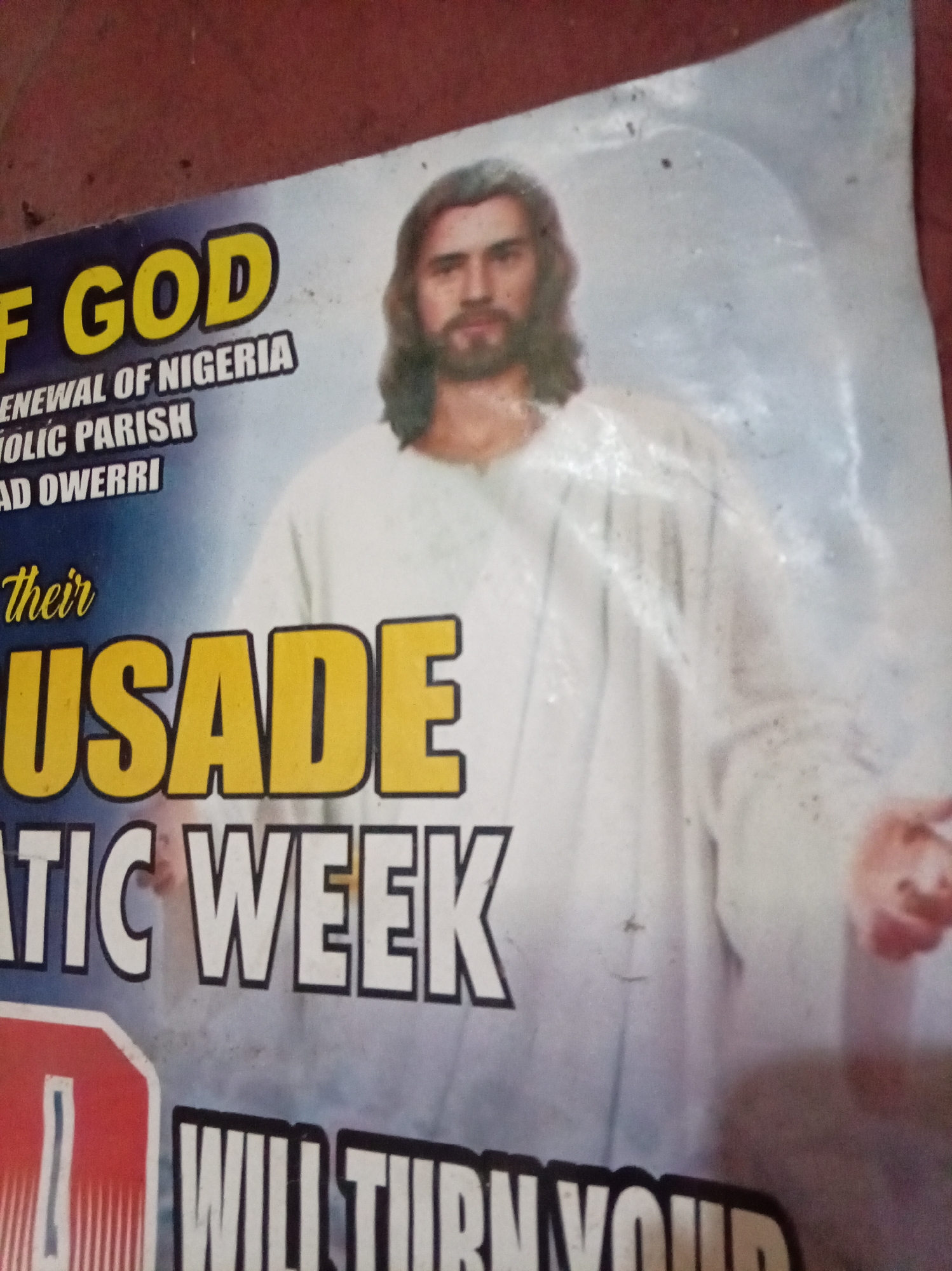 The image shows a poster advertising a Catholic Parish's "Crusade" in Owerri, Nigeria. The poster is partially visible and features a depiction of Jesus Christ wearing a white robe. Above "Crusade" are the words "of God, Renewal of Nigeria Catholic Parish, Ad Owerri." Below "Crusade" are the words "tic Week" and partially visible text that begins with "Will Turn Your." The poster has some stains and imperfections.