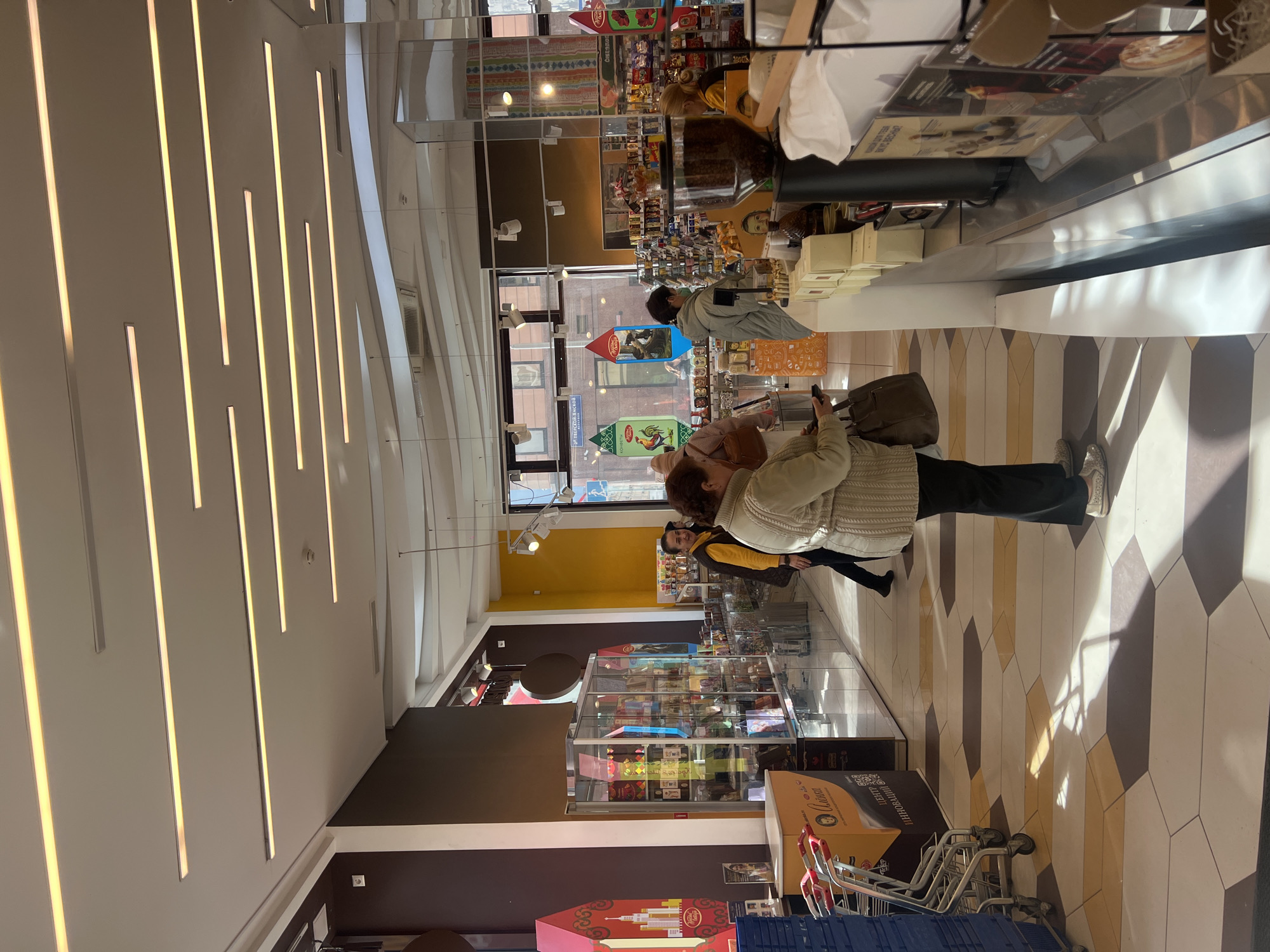 The image captures the interior of a retail store, possibly a confectionery or specialty food shop, located in Moscow, Russia. A woman wearing a cream-colored sweater and dark pants stands in the aisle, holding a mobile phone. Nearby are stacked shopping carts. The store features display cases filled with items. The flooring consists of a geometric pattern of alternating light and dark tiles. The ceiling has recessed lighting fixtures, and the walls are adorned with vertical light strips. Through the store windows, a glimpse of the outside street and building can be seen. Other people are also in the store browsing and shopping.