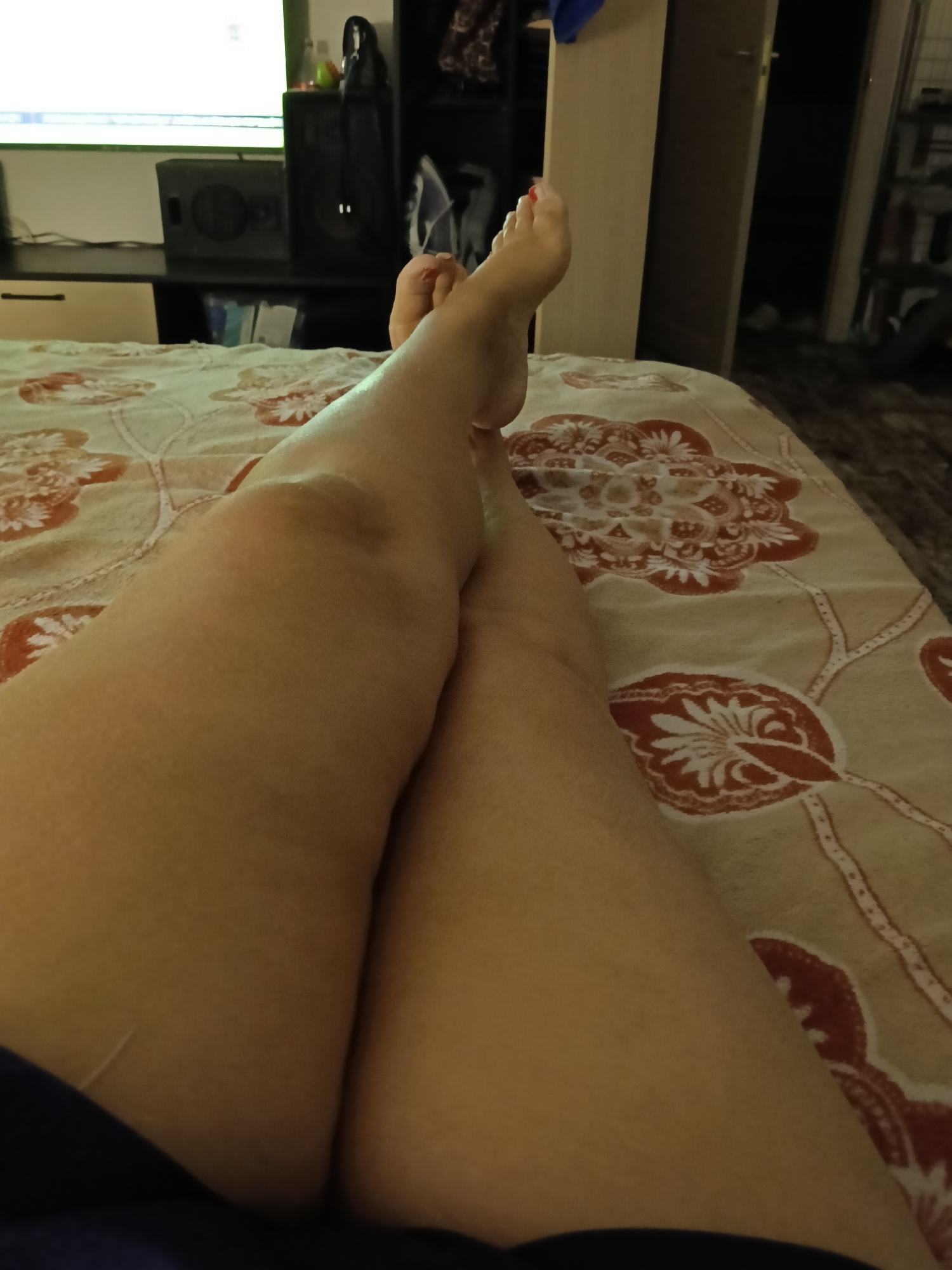 A first-person perspective shows a pair of legs, with the right leg crossed over the left, resting on a bed with a floral-patterned comforter. The feet are visible, with the right foot showing red nail polish. In the background, out of focus, a television screen displays a bright image, and a black speaker is visible on a shelf. To the right, a dark wardrobe door is slightly ajar, revealing a sliver of a dimly lit interior. The setting appears to be a bedroom in a residential apartment in Moscow, Russia.