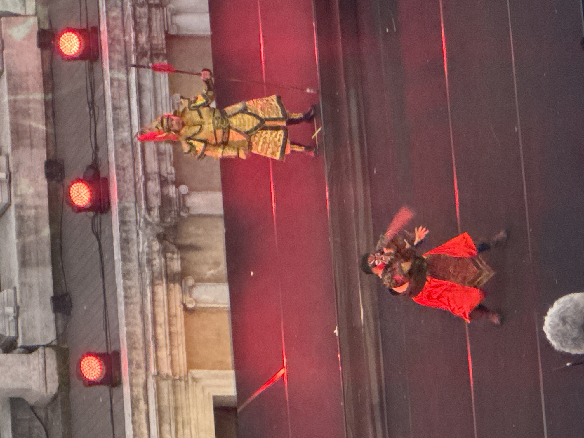 The image depicts an outdoor performance at the Ancient Roman Theatre in Plovdiv, Bulgaria, illuminated by red stage lights. In the foreground, a performer in a red and gold costume with a theatrical mask is in motion, possibly engaged in a fight scene. In the background, another performer stands in a detailed gold and yellow armor, holding a spear with a red flag. The theatre's ancient architecture is visible in the background, with stone columns and ledges. The scene appears to be a historical reenactment or a theatrical play.