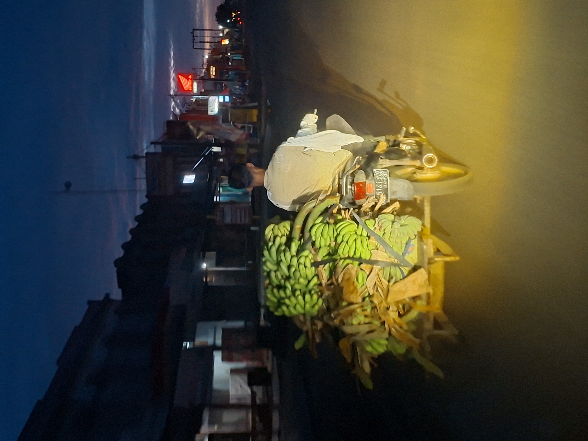 An elevated rear-view photograph captures a male individual operating a motorcycle with an attached sidecar on an asphalt road at twilight or dawn in Grong-Grong, Indonesia. The rider, observed from behind, is wearing a light-colored long-sleeved shirt. The motorcycle displays a license plate reading '5141 NM 08.22'. The sidecar is heavily laden with numerous bunches of unripe green bananas and dried plant material, secured by visible restraints. The immediate foreground of the road is brightly illuminated by a strong yellow light source, casting elongated shadows of the vehicle and rider. The sky above is dark blue, featuring scattered clouds with subtle reddish-purple twilight hues. Structures consistent with commercial buildings or residences line both sides of the street. On the right side of the road, multiple illuminated signs are visible, including a prominent red Honda logo, a 'BSI ATM' sign, and a sign identifying 'LAXANA MOTOR'. Streetlights are active along the thoroughfare in the distance.