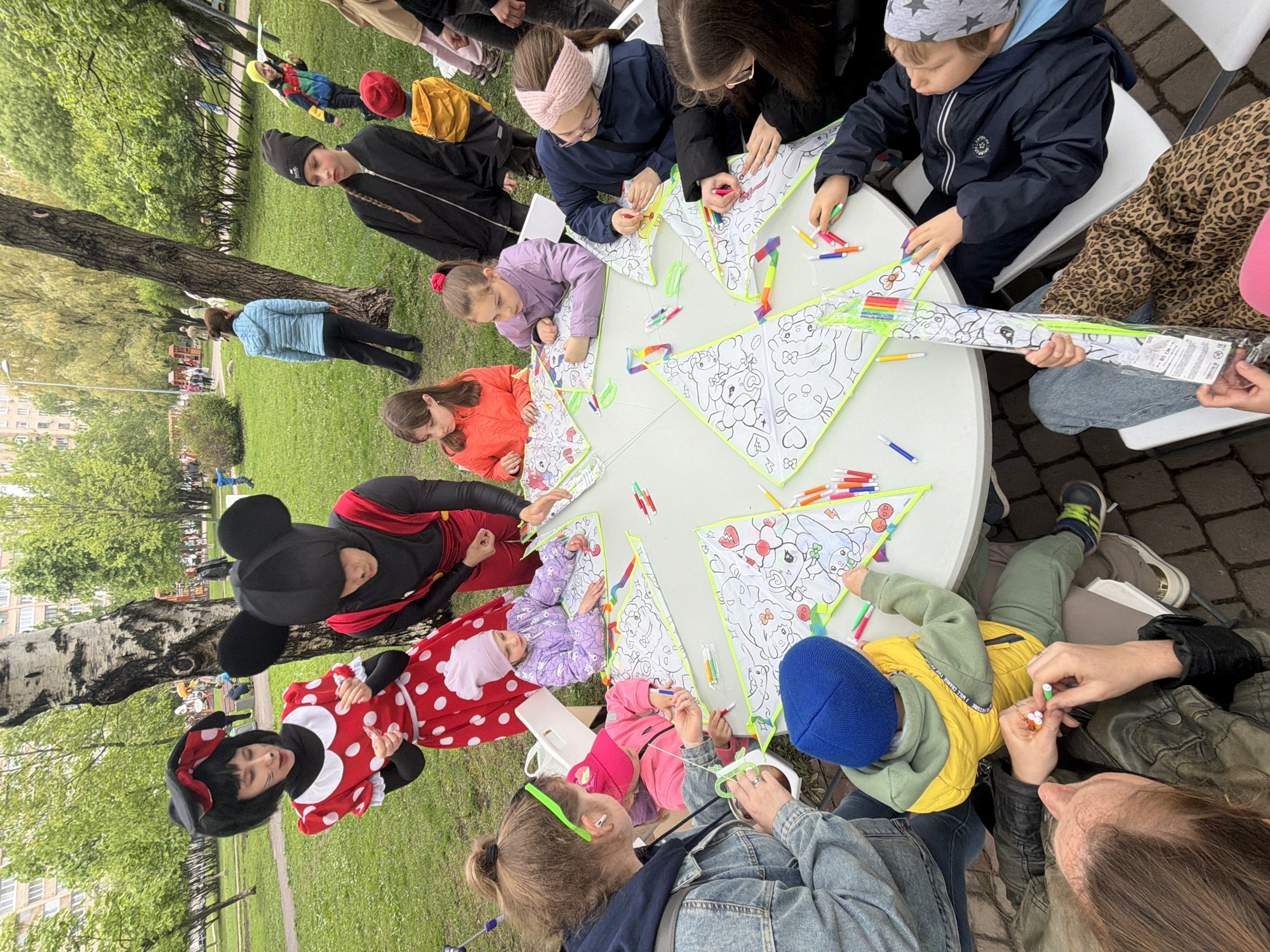 The scene captured is an outdoor event in a park, likely in the city of Saint Petersburg, Russia, given the image title "Saint Petersburg, Russia, June 2024." A group of children and adults are gathered around a large, round, white table, engaged in coloring triangular kites, which are mostly white with printed cartoon outlines. Numerous colorful markers are scattered across the tabletop. The participants include children of various ages, some wearing hats or headbands, focusing on their artistic endeavors. Adults are assisting the children. Two individuals dressed as Mickey and Minnie Mouse are visible near the table, likely part of the event's entertainment. The setting is a park with grass and trees in the background. The ground surrounding the table is partially bricked.
