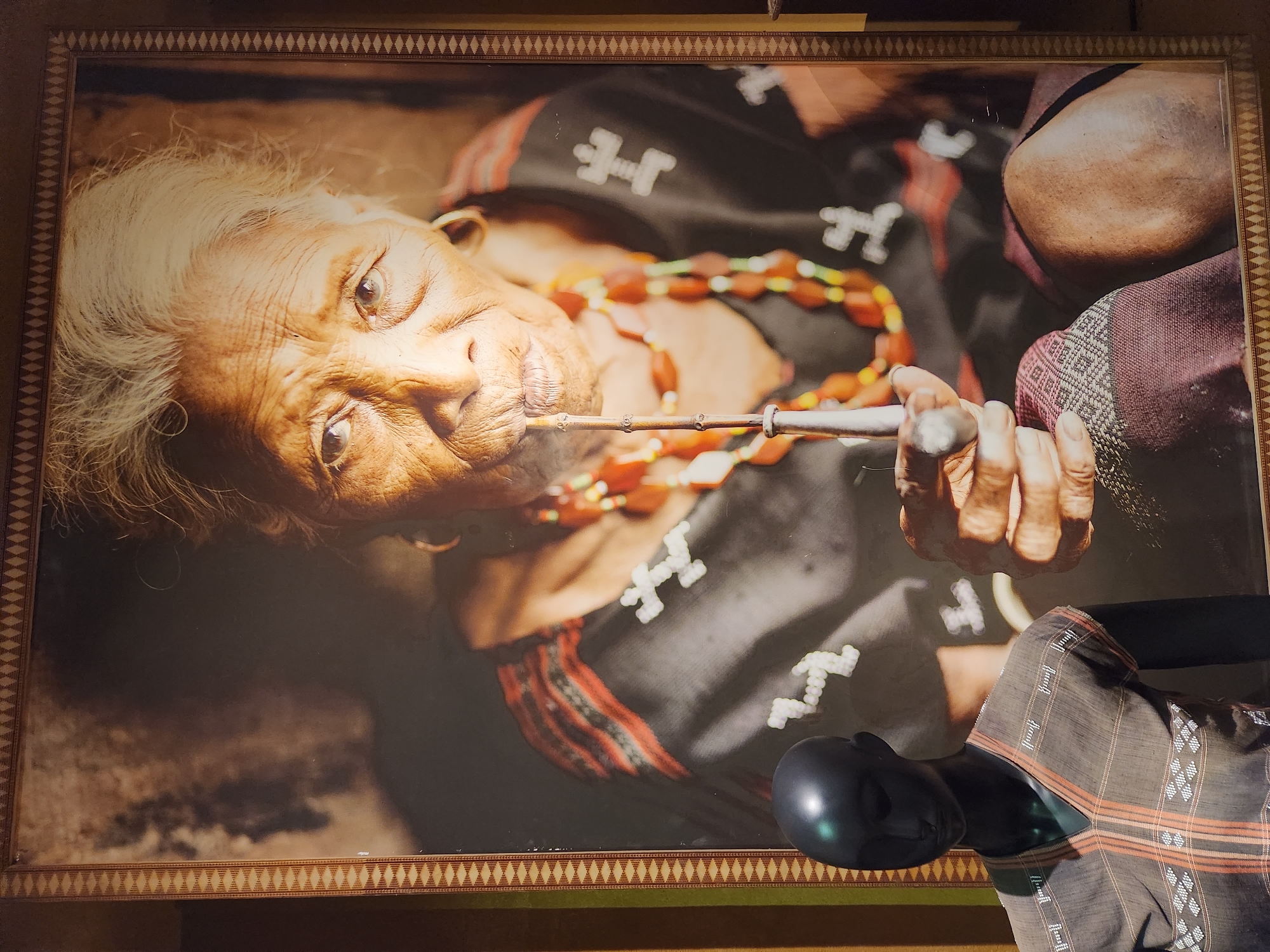 This media file features a framed photograph of an elderly person, likely a woman, in what appears to be a cultural setting in Hội An Ward, Vietnam. The subject is depicted from a high-angle shot, lying down, with their head tilted towards the viewer. They have white hair and deeply wrinkled skin, suggesting advanced age. The person is wearing a dark garment with intricate patterns, including white cross-like symbols and red and black striped bands. A prominent feature is a necklace made of segmented, reddish-orange beads.
The subject is holding a long, thin object with both hands, bringing it to their mouth, suggesting they are smoking a pipe or a similar implement. Wisps of smoke are visible rising from the object. The person's expression is intense and direct, with their eyes looking towards the camera. The background is dark and blurred, focusing all attention on the subject and the activity.
A portion of a mannequin dressed in a patterned shirt is visible in the lower right corner of the image, slightly obscuring the bottom edge of the framed photograph. The frame itself has a decorative border with a repeating diamond pattern. The lighting suggests an indoor setting, possibly a display or a home. The overall mood is one of contemplation and perhaps resilience, captured in a close-up portrait. There is no visible text in the image.
