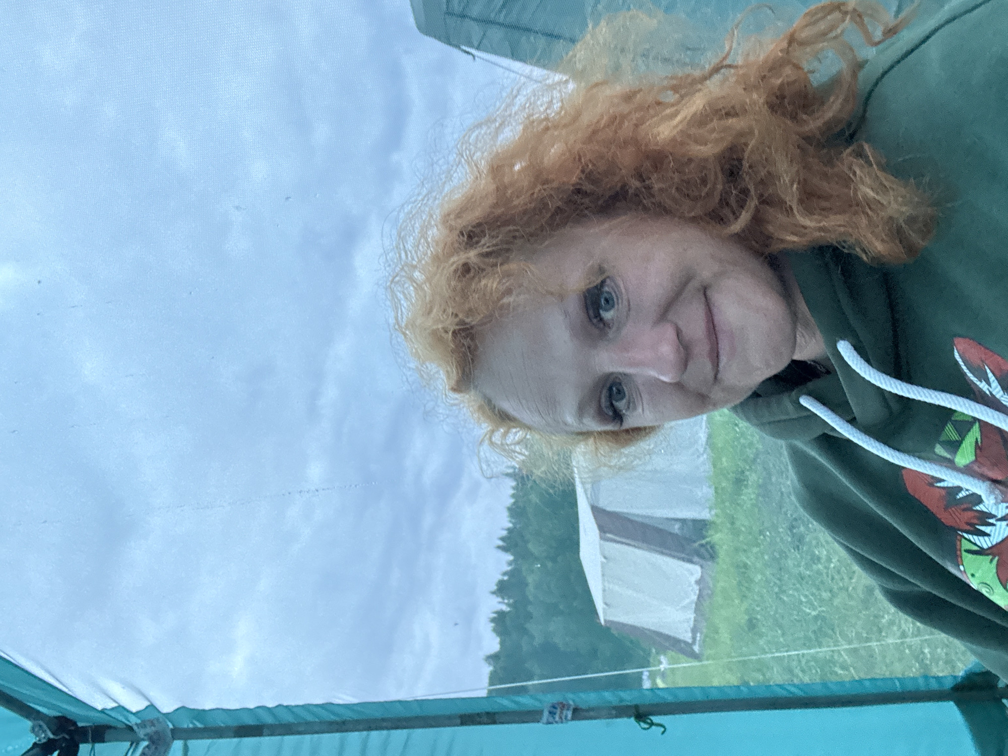 A woman with voluminous, red, curly hair is depicted in a front-facing portrait photograph, taken from inside a structure and looking outward. She has light-colored eyes and a subtle smile, wearing a dark green hooded sweatshirt with white drawstrings and a multi-colored graphic design on the chest. The image is captured through a translucent screen or panel, framed on the left by a teal structural element. Beyond this screen, a grassy field is visible, featuring two tents in the mid-distance: a larger, light-colored tent with a visible A-frame structure, and a smaller, partially obscured dark-colored tent to the right. A dense line of green coniferous trees forms the horizon under an overcast, light blue-gray sky. This scene is located in Bolshaya Kivara, Russia.