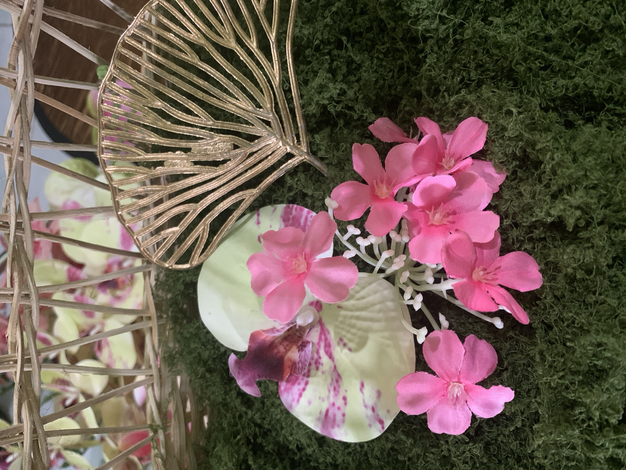 This is a close-up, high-angle shot of a decorative arrangement. In the foreground, a bed of textured green material resembling moss serves as the base. Scattered across the moss are several bright pink, five-petaled flowers, some with white stamen. To the left, a large pale green orchid petal with pink speckles is visible, with a dark purple and pink center. Behind the orchid, a gold-colored, intricate metal leaf-shaped ornament is positioned. In the upper background, parts of a woven basket made of light-brown reeds are visible, with blurred yellow and pink floral elements behind it. The lighting is soft, and there are no people or animals present. The scene appears to be an indoor decorative display, with no discernible connection to Kuala Pembuang, Indonesia.
