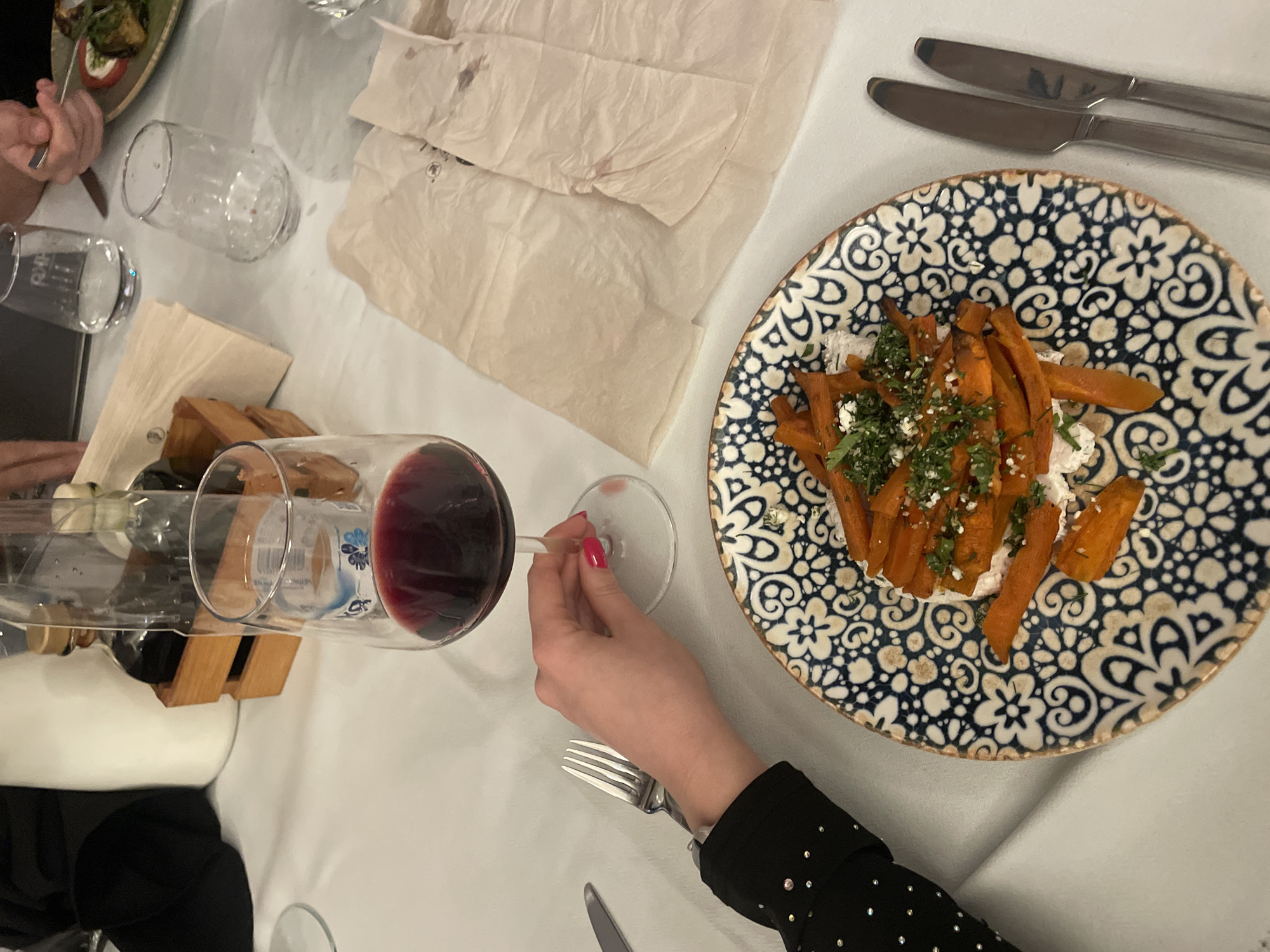 The image depicts a table setting at a restaurant, possibly located in Varna, Bulgaria, as indicated by the Cyrillic text on a bottle label. A person is holding a glass of red wine. A plate with a blue and white pattern contains orange-colored food, possibly sweet potato fries, topped with herbs and white sauce. There are two knives and a fork placed on the table, along with napkins. A wine holder with bottles is visible in the background, as well as other glasses. The table has a white tablecloth. Another person is also visible near the top left of the frame eating from a plate.