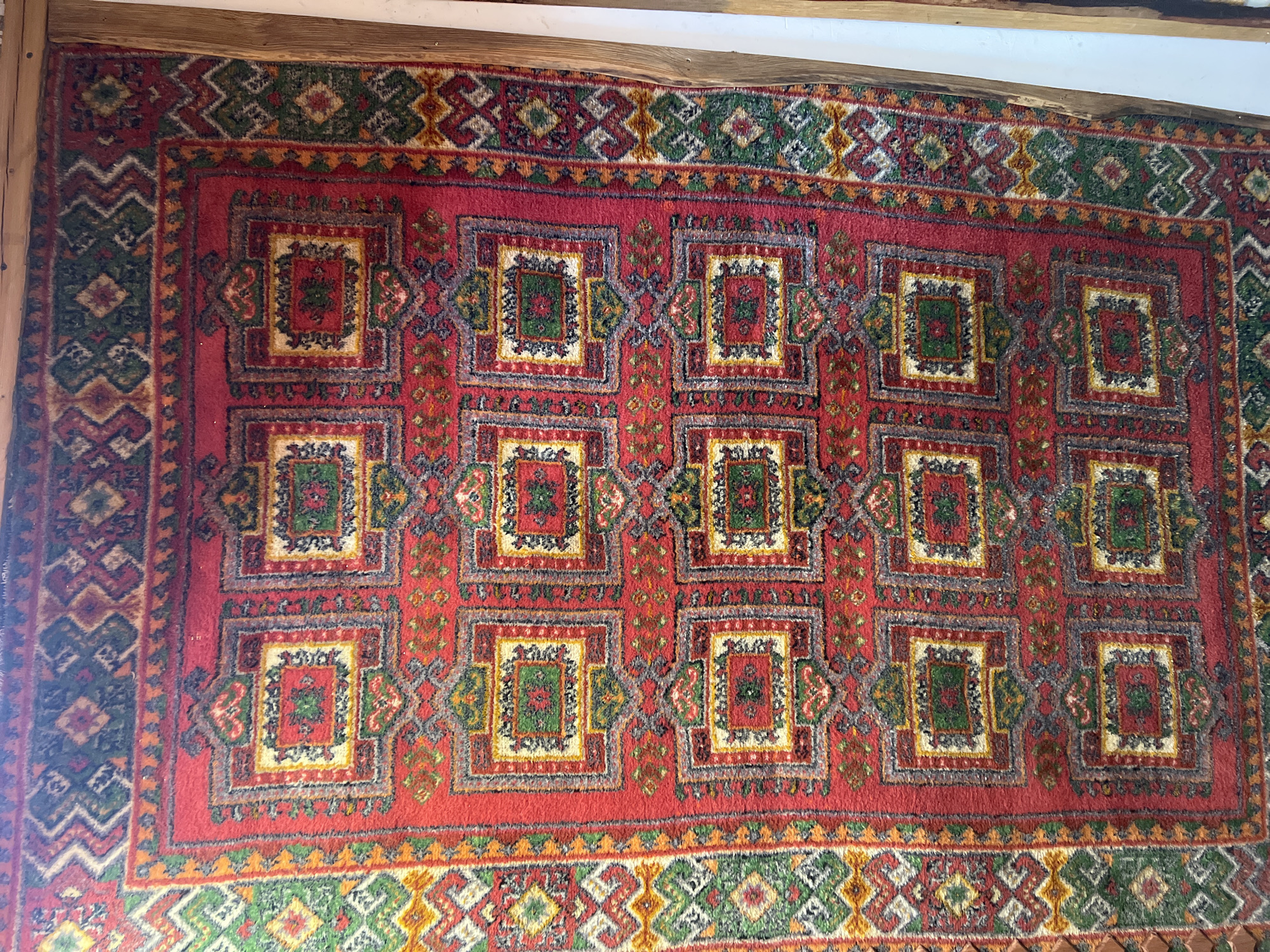 A red, intricately patterned rug is displayed horizontally, filling most of the frame. The rug features a repeating geometric design, with rows of square motifs. Each square contains smaller, nested squares and diamond shapes in shades of green, yellow, white, and grey. The dominant color of the rug is a deep red. Surrounding the central pattern is a border with zig-zag and diamond patterns in green, yellow, and white. The rug is placed on a light-colored surface, likely a floor, and wooden elements are visible at the bottom edge of the image, suggesting it might be part of a display or furniture. The lighting appears to be natural and slightly diffused, casting soft shadows and highlighting the texture of the rug. The location is identified as Norviliškės, Lithuania.
