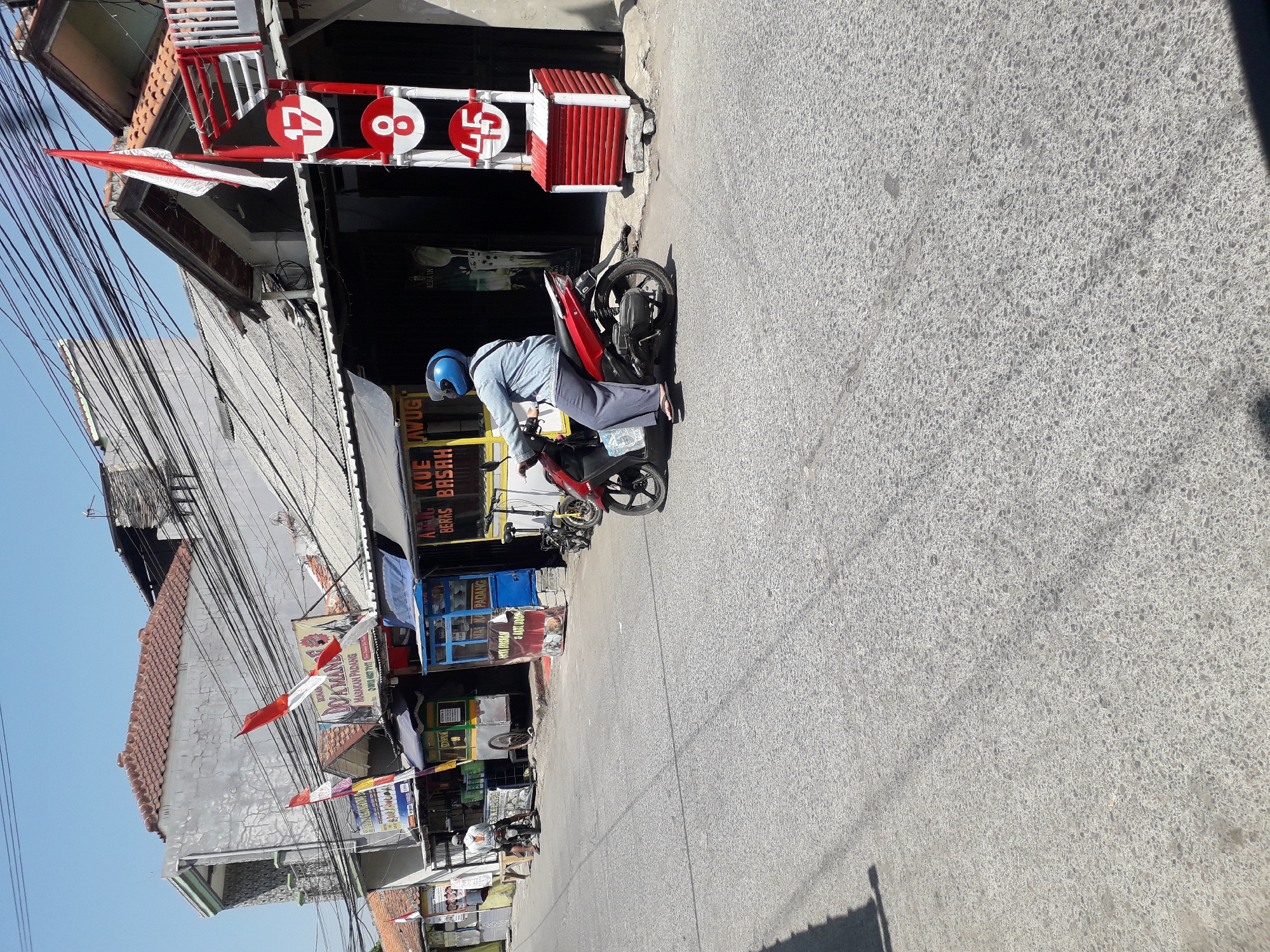 The image captures a street scene in Linggar, Indonesia, on a sunny day. In the foreground, an individual wearing a blue helmet and a light blue long-sleeved shirt is riding a red motorcycle. They are wearing dark pants and flip-flops. The motorcycle has a white plastic bag hanging from its rear.
The background features several buildings with corrugated metal roofs and some older tiled roofs. Numerous electrical wires crisscross overhead. Along the side of the street, there are various small shops and food stalls. One stall displays a sign that reads "RUMAH MAKAN PADANG" and another has "KUE KULIT" visible. To the right, a red and white vertical sign has circles with numbers: "17", "8", and "45". Indonesian flags are visible attached to poles and buildings, suggesting a national holiday or celebration.
The road surface is asphalt with visible texture and some scattered debris. The overall environment appears to be a busy, commercial area within a residential neighborhood.