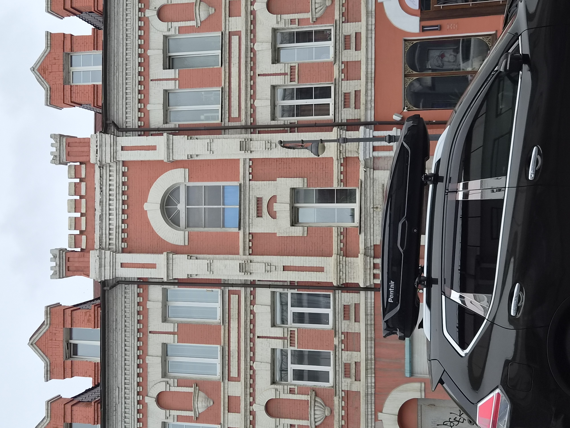 A black SUV with a black rooftop cargo carrier is parked in front of a multi-story building with a salmon-colored facade and white decorative trim. The building has a symmetrical design with numerous windows. A lamppost stands to the right of the vehicle. The scene is likely located in Vladivostok, Russia, given the architectural style.