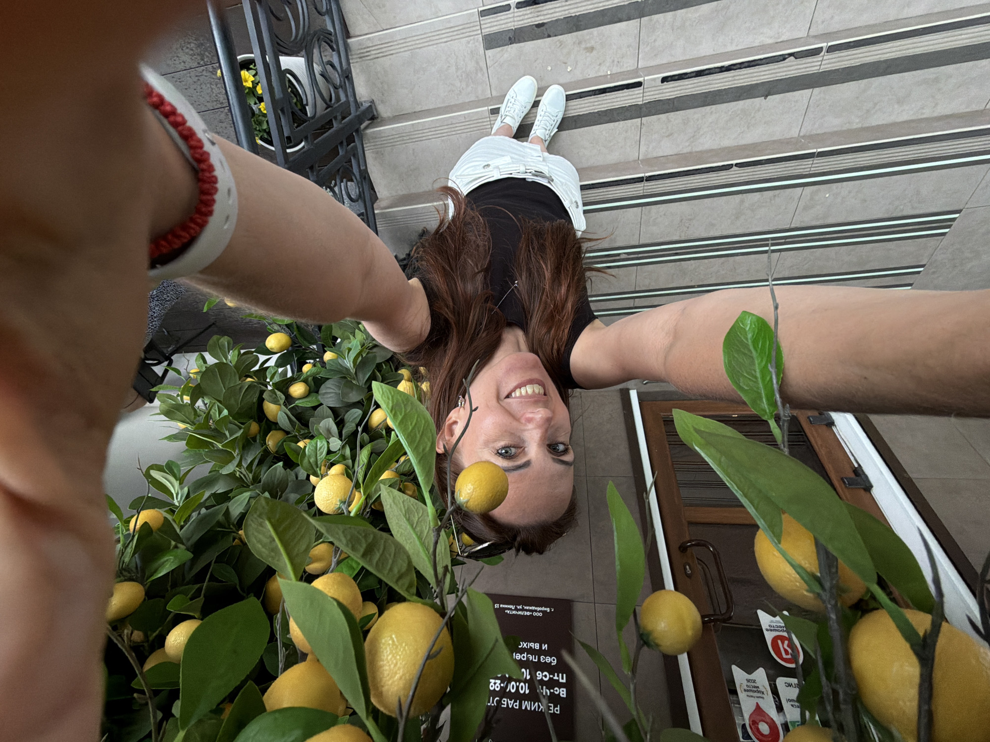 A woman takes a selfie in an outdoor setting surrounded by artificial lemons and green leaves. She is wearing a black sleeveless top, a white skirt, and white sneakers. The background shows a building with a sign in Russian, indicating the location as Birobidzhan, Russia. The sign contains text about working hours and an address. The floor is tiled with a patterned design. The woman is smiling and looking at the camera.
