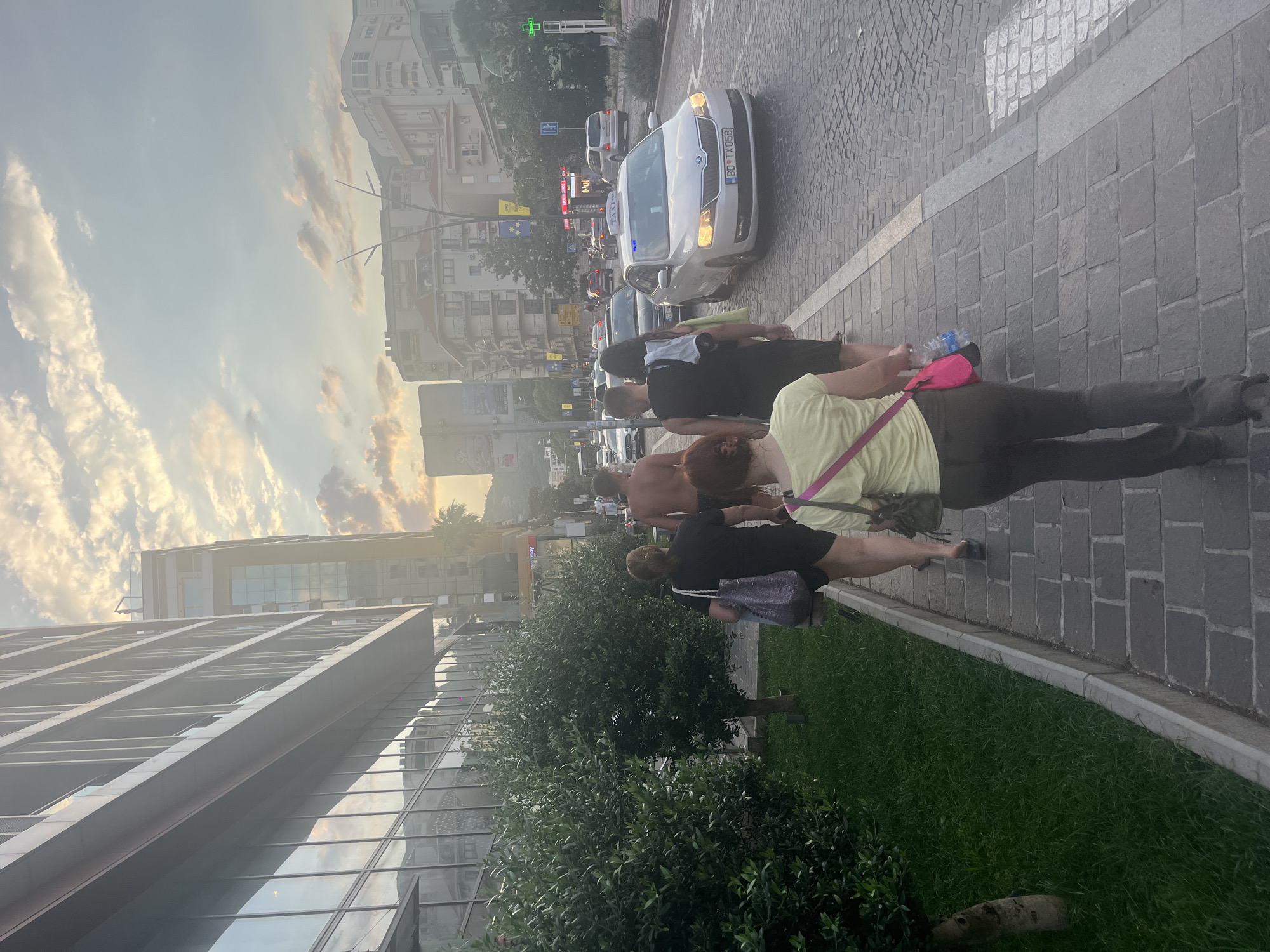 A group of five people walks along a paved sidewalk next to a row of green bushes and a modern building with reflective windows. The sky above is filled with dramatic clouds and the setting sun casts a warm glow on the scene. A man in the group is shirtless. A white taxi with a "TAXI" sign on its roof is driving on the street in the background, along with other cars and a pedestrian crossing. Buildings and trees line the street, suggesting an urban environment.