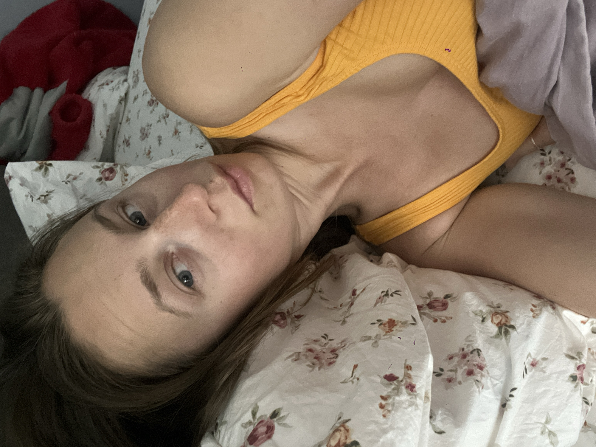 A forensic description of the scene in Bečići, Montenegro, reveals an indoor setting, presumably a bedroom. A single female individual, appearing to be an adult, is depicted lying in a bed. Her head rests on a white pillow adorned with a floral pattern featuring pink, red, and green elements. The woman has light-colored skin, light brown hair, and blue eyes. Visible freckles are present on her face, particularly across her nose and cheeks. She is looking directly at the camera. She wears a ribbed, yellow sleeveless top with a scoop neckline. A light grey fabric, possibly a blanket or duvet, partially covers her lower torso and left arm. In the upper right background, a piece of red fabric, possibly clothing or a blanket, is visible atop another white item with a floral pattern, consistent with bedding. The overall lighting suggests an interior environment.