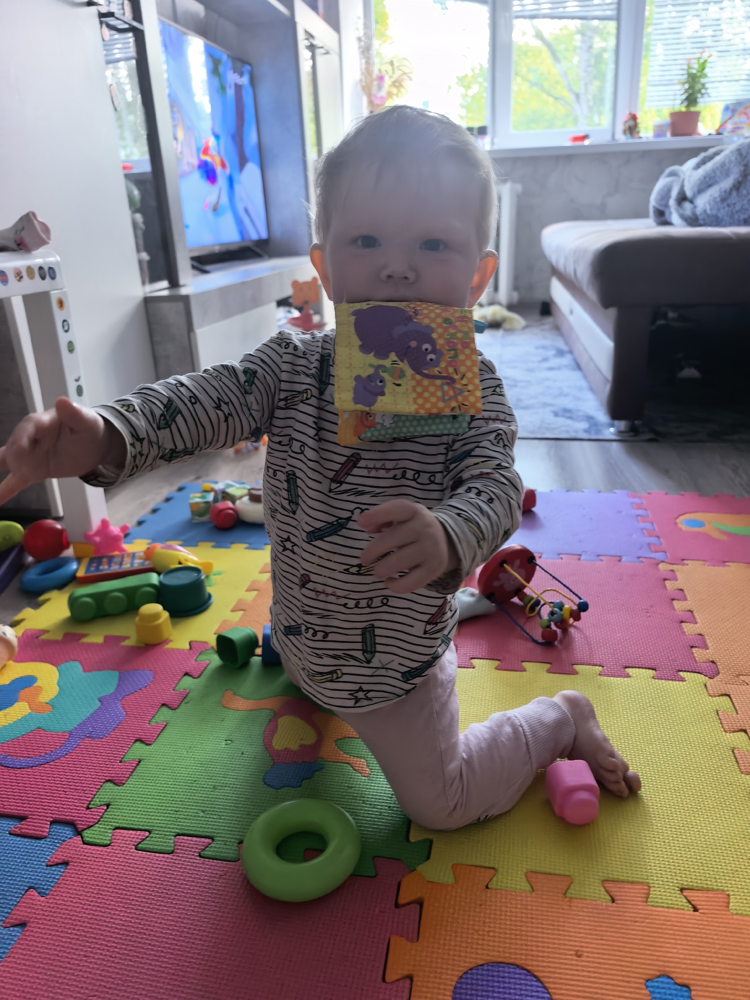 A toddler is kneeling on a colorful foam play mat in a room. The child is wearing a striped long-sleeved shirt and pink pants and is holding a fabric block in their mouth. The room is furnished with a TV, a couch, and a white shelving unit. Toys are scattered on the play mat. The location is Sterlitamak, Russia.