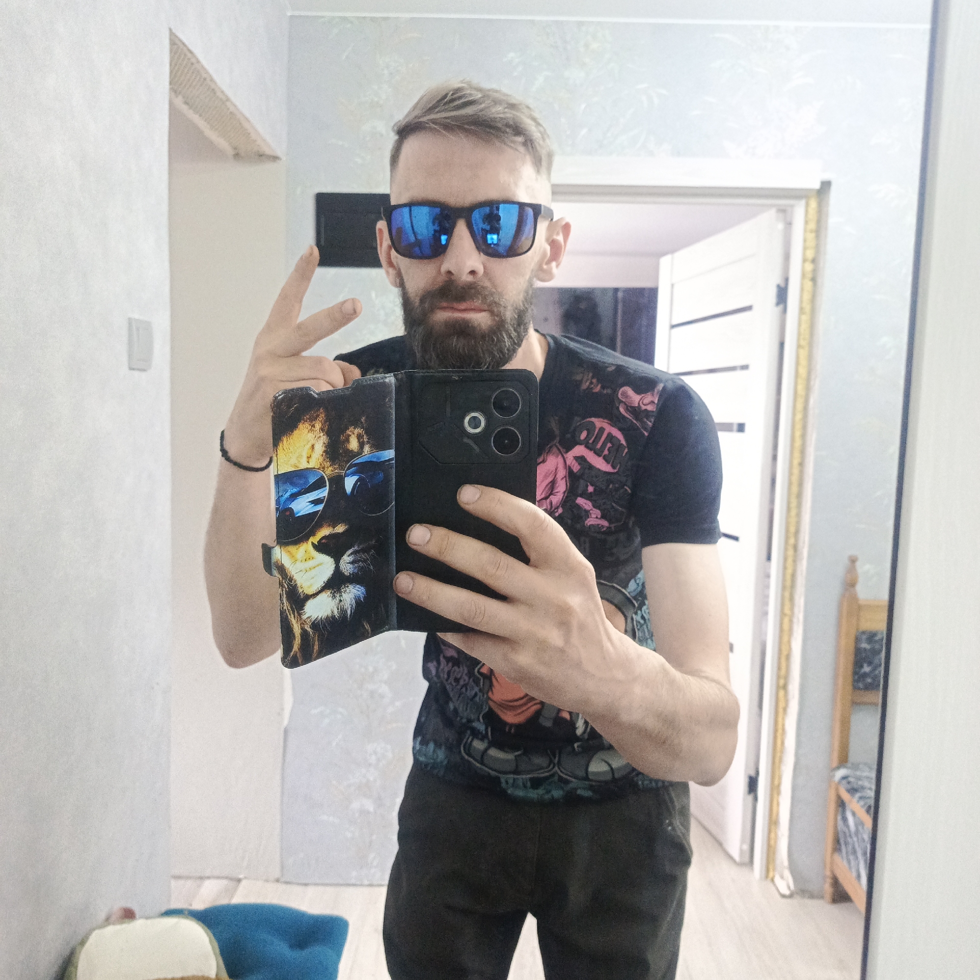A man with a beard and sunglasses is taking a selfie in a hallway. He is holding a phone with a lion design case. The hallway has light-colored walls and a doorway leading to another room, which contains a bed. The reflection in the mirror shows a bed with blue and white bedding. The precise location in the city of Марковский, Russia cannot be confirmed from the image alone.