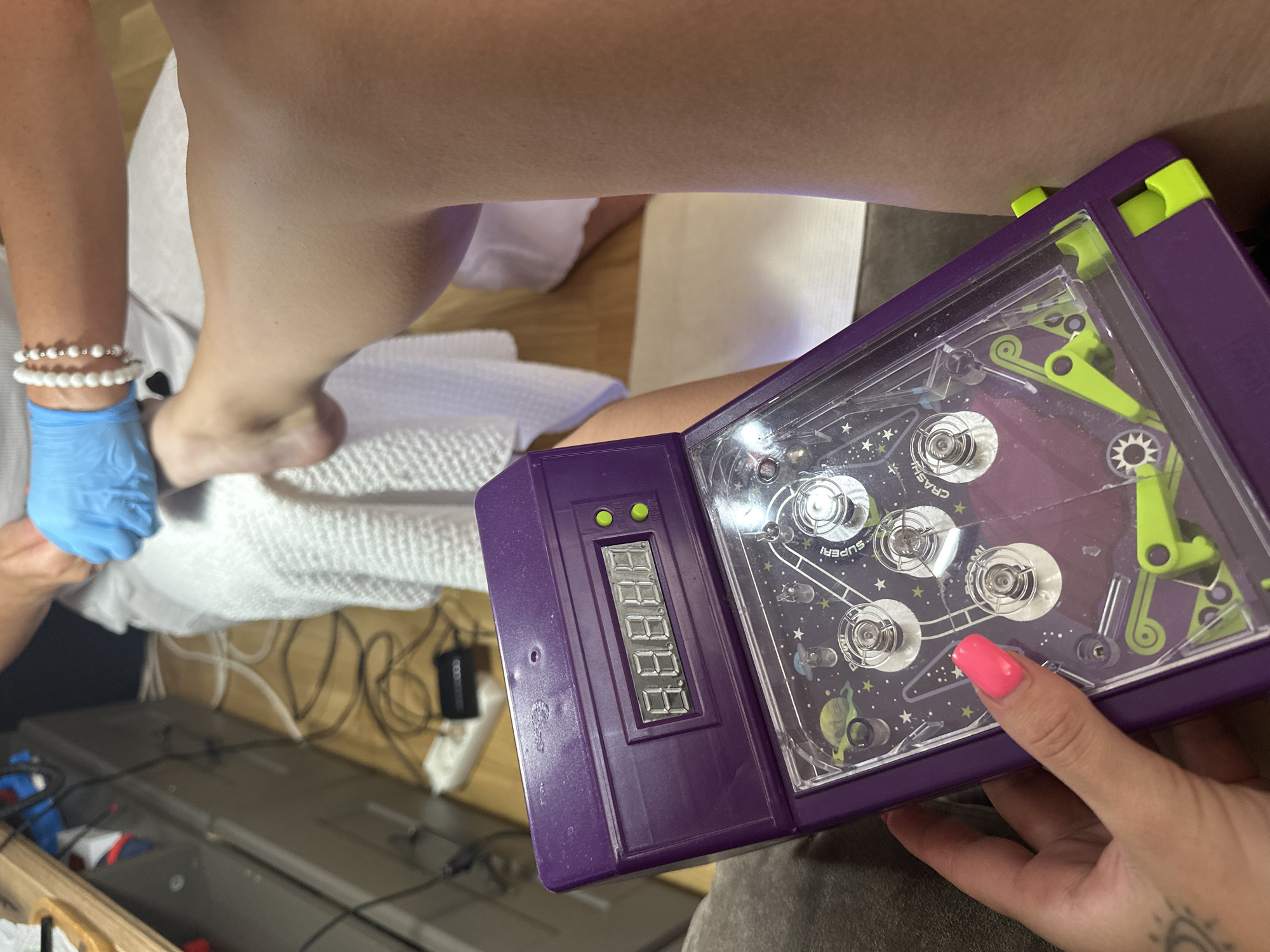The image shows a close-up of a person holding a purple electronic pinball game. The game has a digital display that shows "8.8.8.8.8". The pinball machine has a space-themed playfield with planets and stars. The person's hand is visible, with a bright pink fingernail. In the background, a person wearing blue gloves is attending to a foot, which is covered by a white towel. The floor is visible, and appears to be made of wood. There are electrical cords and a power strip on the floor. The scene is indoors, likely in a salon or home setting, with no identifiable landmarks to suggest a specific location within Budva, Montenegro.