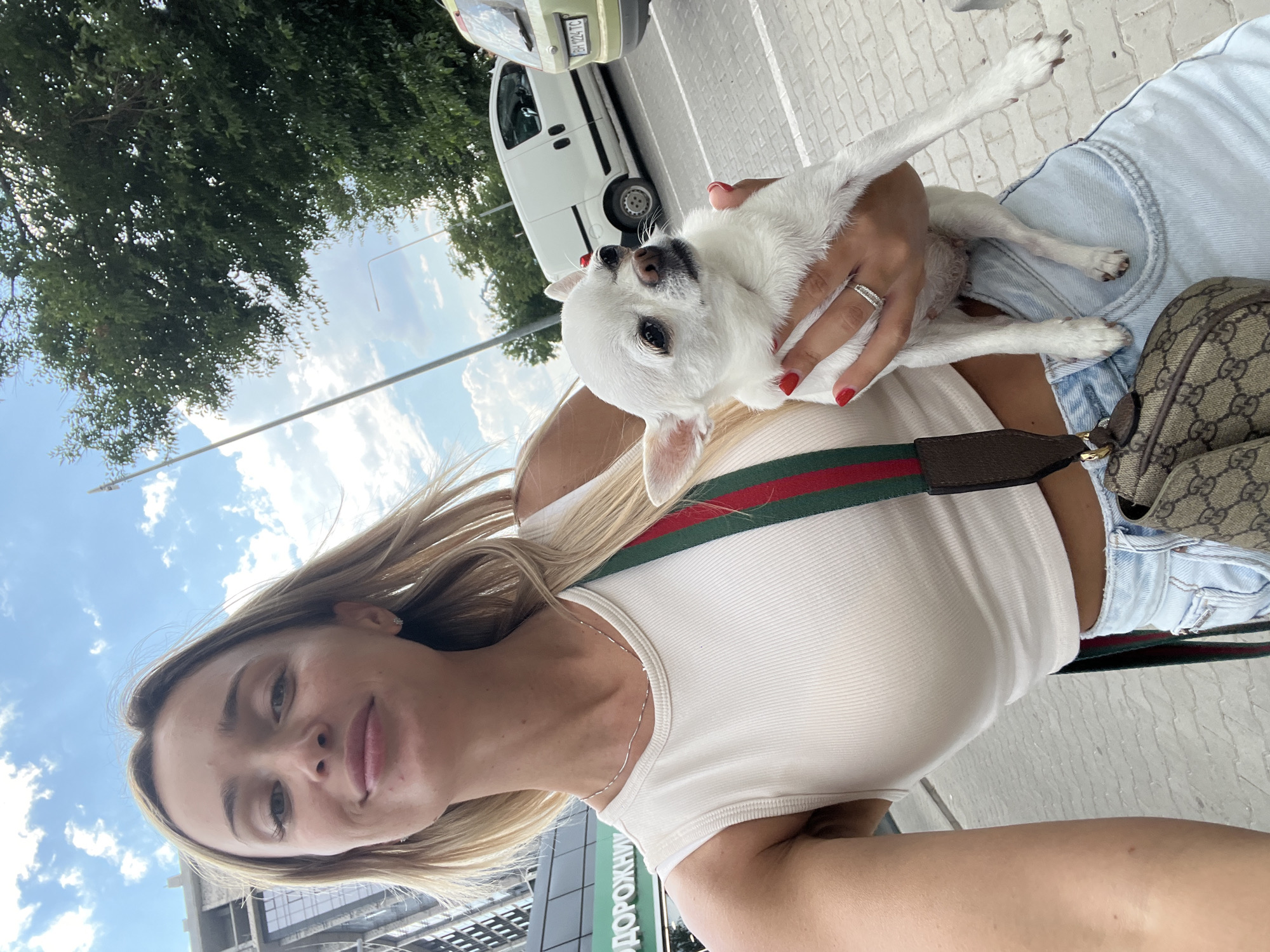 A woman with blonde hair is holding a small white chihuahua dog. She is wearing a white tank top and denim shorts. A Gucci bag is visible on her hip. The background shows a street scene with a white van, a light-colored car, trees, and a building. The sky is blue with some clouds. The location is Fontanka, Ukraine.