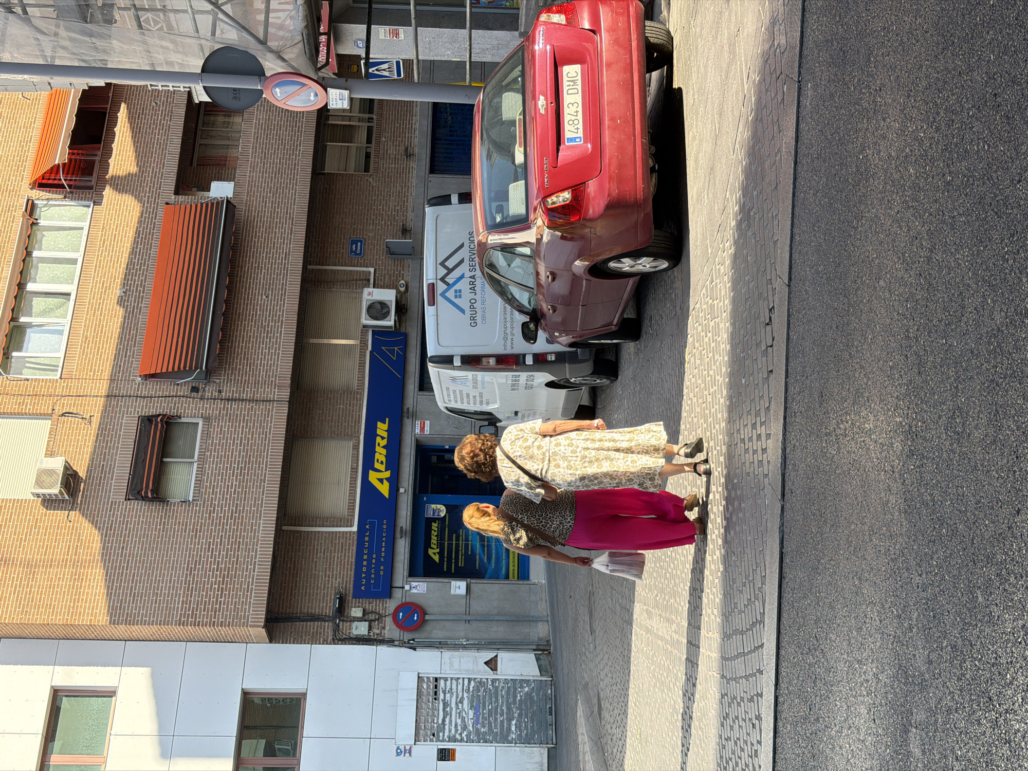 Two women are walking on a sidewalk in front of a brick building. The woman on the left is wearing pink pants and a leopard-print top. The woman on the right is wearing a floral-print dress. Both women are walking away from the camera. To the right of the women, there is a red Chevrolet sedan with the license plate "4843 DMC". Next to the car is a white van with "GRUPO JARA SERVICIOS" written on the side. The building behind them has a blue awning with the word "ABRIL" in yellow letters. The street is paved with cobblestones. The scene appears to be a commercial or residential area. The overall setting is a street in Las Rozas de Madrid, Spain, with buildings and vehicles characteristic of a Spanish urban environment.