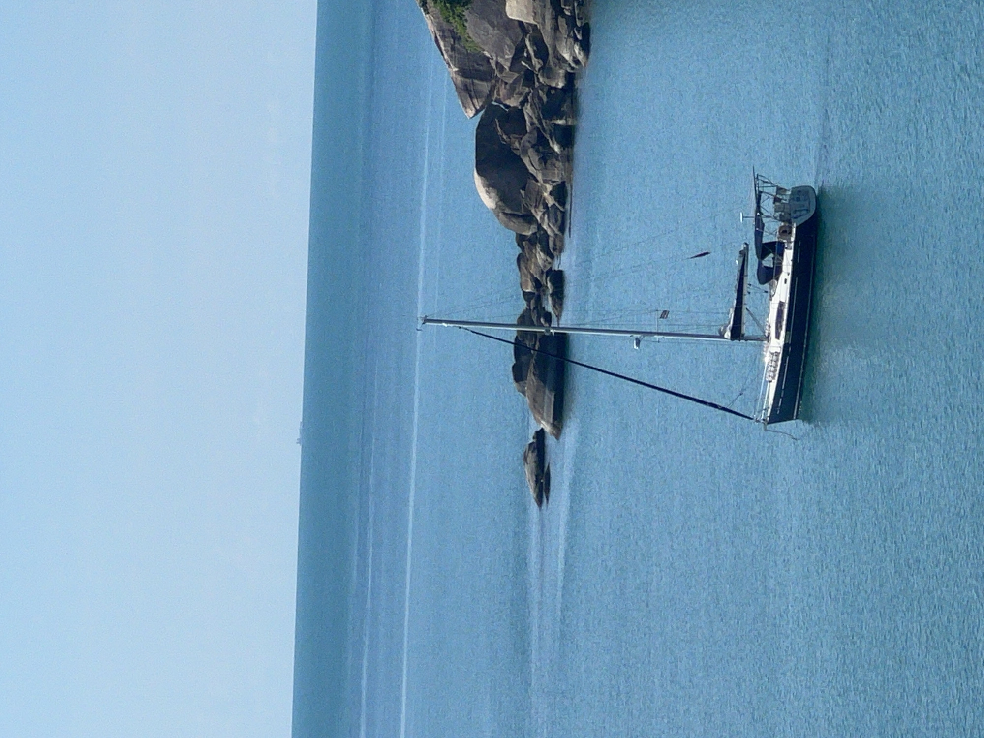 A sailboat is anchored in calm, turquoise water near a rocky coastline under a clear blue sky. The sailboat has a dark hull, white superstructure, and a single mast with sails furled. Several large, rounded boulders are visible in the water close to the shore, and a more extensive rocky outcrop with sparse green vegetation is situated further along the coast. In the far distance, a small, indistinct ship is visible on the horizon. The image captures a serene coastal scene in Baan Chaweng Noi, Thailand.
