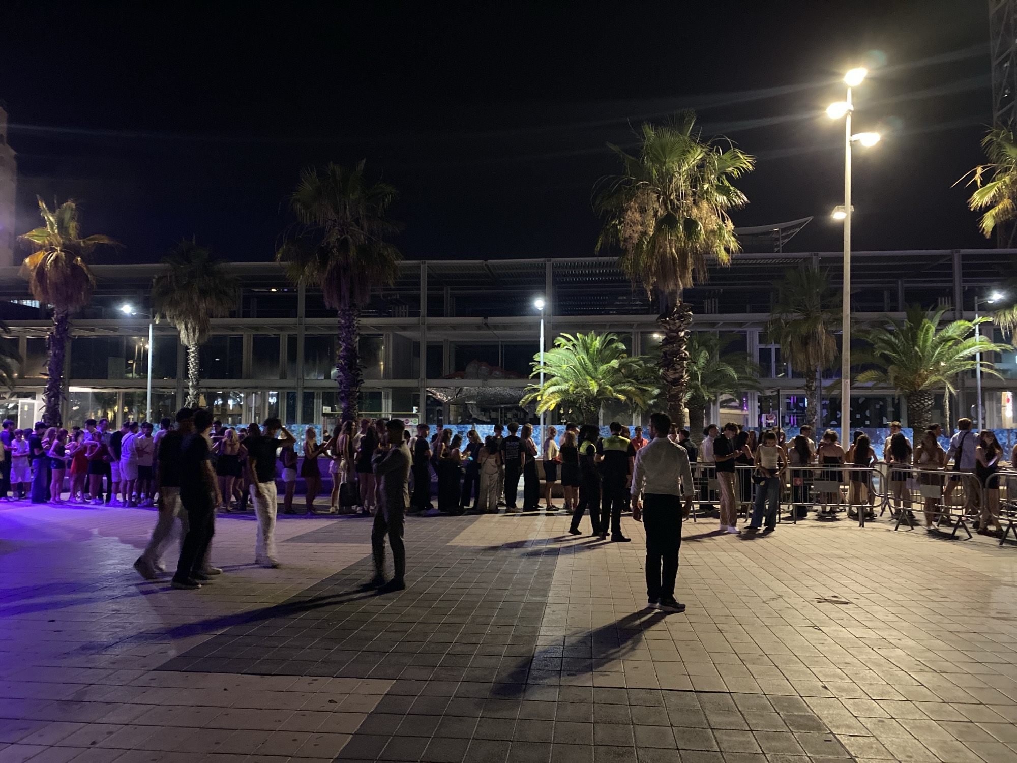 The image depicts a night scene in Barcelona, Spain, likely on a promenade or waterfront area, characterized by warm evening weather. A group of people are gathered on a paved area with square tiles, illuminated by streetlights and ambient building lights. Palm trees are prominent in the background, suggesting a Mediterranean setting.
In the foreground, several individuals are walking or standing. One person in a white shirt and black pants is facing away from the camera, with their shadow cast on the ground. To their left, a group of people are walking, some with their backs to the viewer, others facing sideways.
Further into the scene, a crowd of people are queuing behind a metal barrier, facing a building or an event. Many are dressed in casual summer attire, with women in dresses and men in t-shirts and shorts. Some individuals in the crowd appear to be holding drinks.
In the middle ground, several more palm trees are visible, some illuminated with purple light from below. A tall lamppost with multiple lights stands to the right of center. The background reveals the facade of a modern building with a skeletal structure and open sections, suggesting construction or an unfinished area. The overall atmosphere appears lively and social, typical of an evening gathering in a public space.