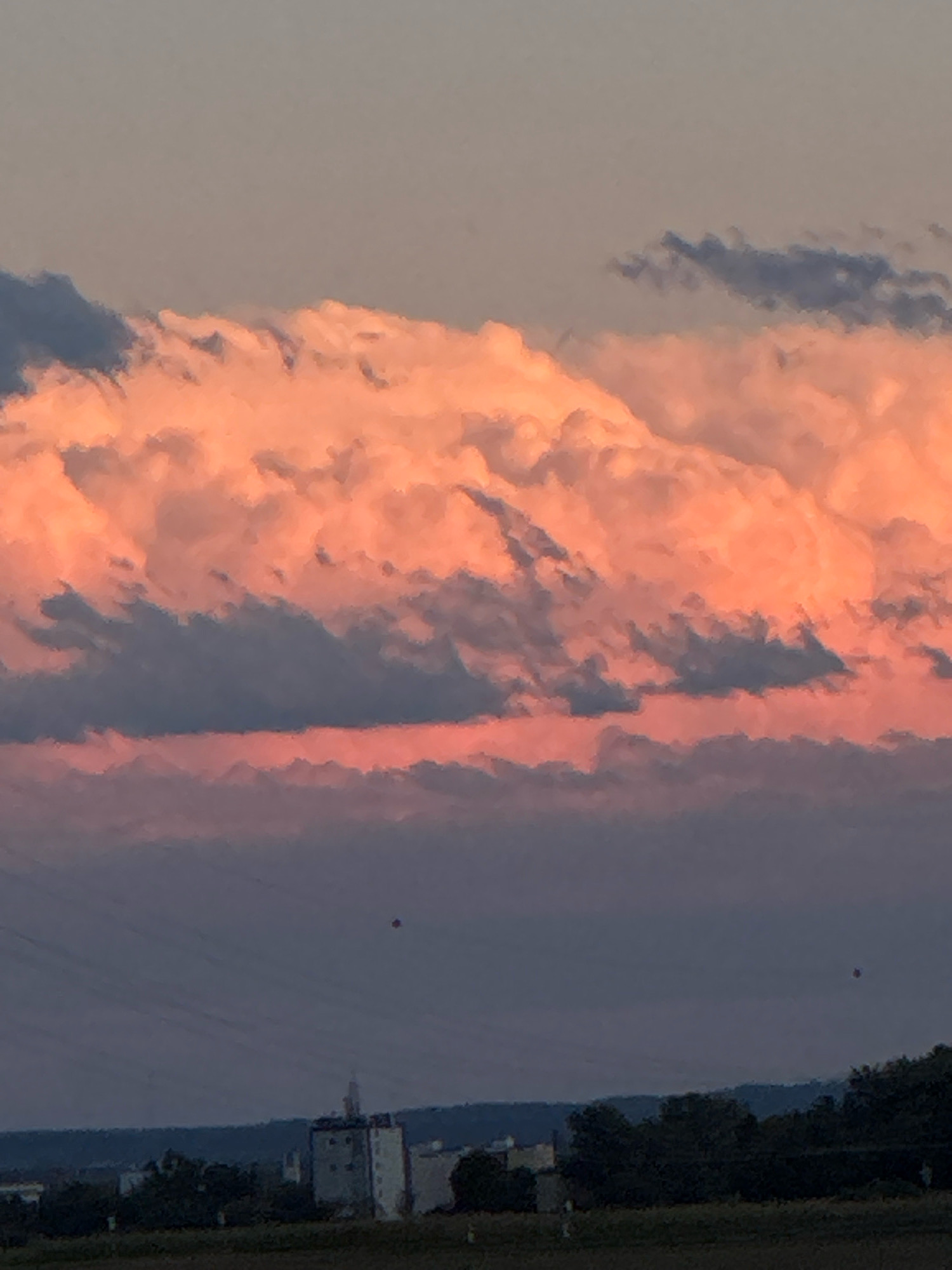 The image shows a sunset over Wettstetten, Germany. The sky is filled with large, fluffy clouds that are illuminated by the setting sun, casting a warm, orange glow. The lower part of the sky is a deep purple, blending into the greyish-blue of the distant horizon. In the foreground, the silhouette of trees and buildings is visible against the colorful sky. There are two small, dark, circular objects in the sky, possibly balloons or drones. The overall impression is one of a peaceful and serene evening.