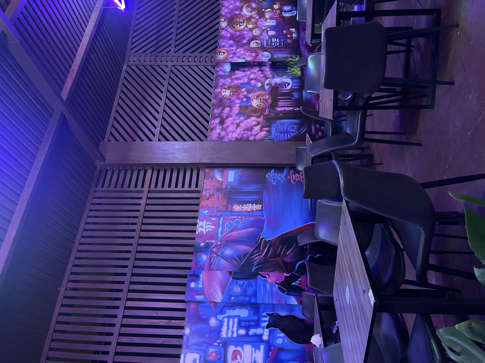 The image shows the interior of a dining establishment in Arica, Chile. The space features a combination of wooden slat walls and mural art. The murals depict Japanese-themed imagery, including cherry blossoms, lanterns, and a figure with an umbrella in a cityscape reminiscent of Tokyo.
The furniture consists of dark chairs with square backs and dark-topped tables. The tables and chairs are arranged in rows, suggesting a restaurant or bar setup. The lighting is predominantly purple and blue.