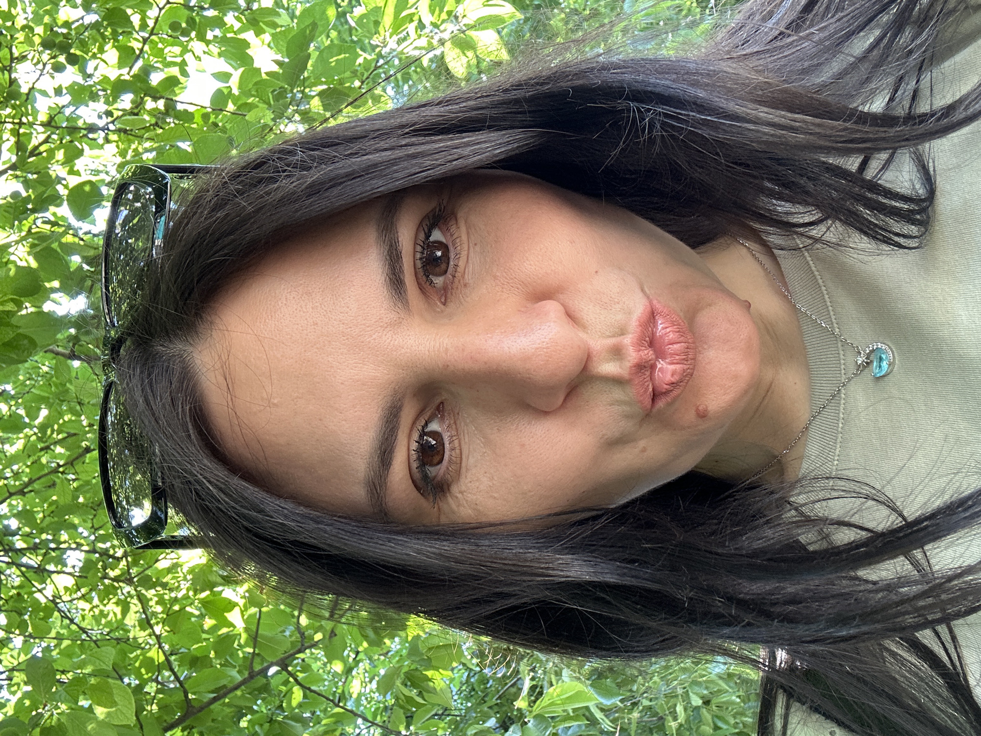 A medium shot portrait of a woman with dark brown hair pulled back by black sunglasses perched on her head, looking directly at the camera with her lips puckered. She is wearing a light green t-shirt and a silver necklace with a crescent moon pendant featuring a blue stone and small clear stones. The background consists of lush green foliage, suggesting an outdoor setting, possibly in a park or garden. The lighting is natural and bright, indicating it is daytime. There are no other individuals present in the image.