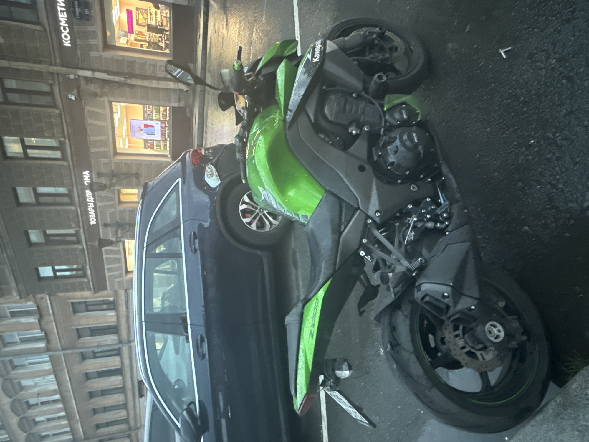 The image captures a Kawasaki Z1000 motorcycle parked on a wet street in Saint Petersburg, Russia. The motorcycle is predominantly green and black. Behind the motorcycle, a dark blue station wagon is parked. In the background, a building with multiple windows and architectural details is visible, with storefronts displaying signage in Russian, including "ТОВАРЫ ДЛЯ ДОМА" (Goods for Home) and "КОСМЕТИКИ" (Cosmetics). The street surface is wet, indicating recent rain. The overall lighting suggests an overcast day.