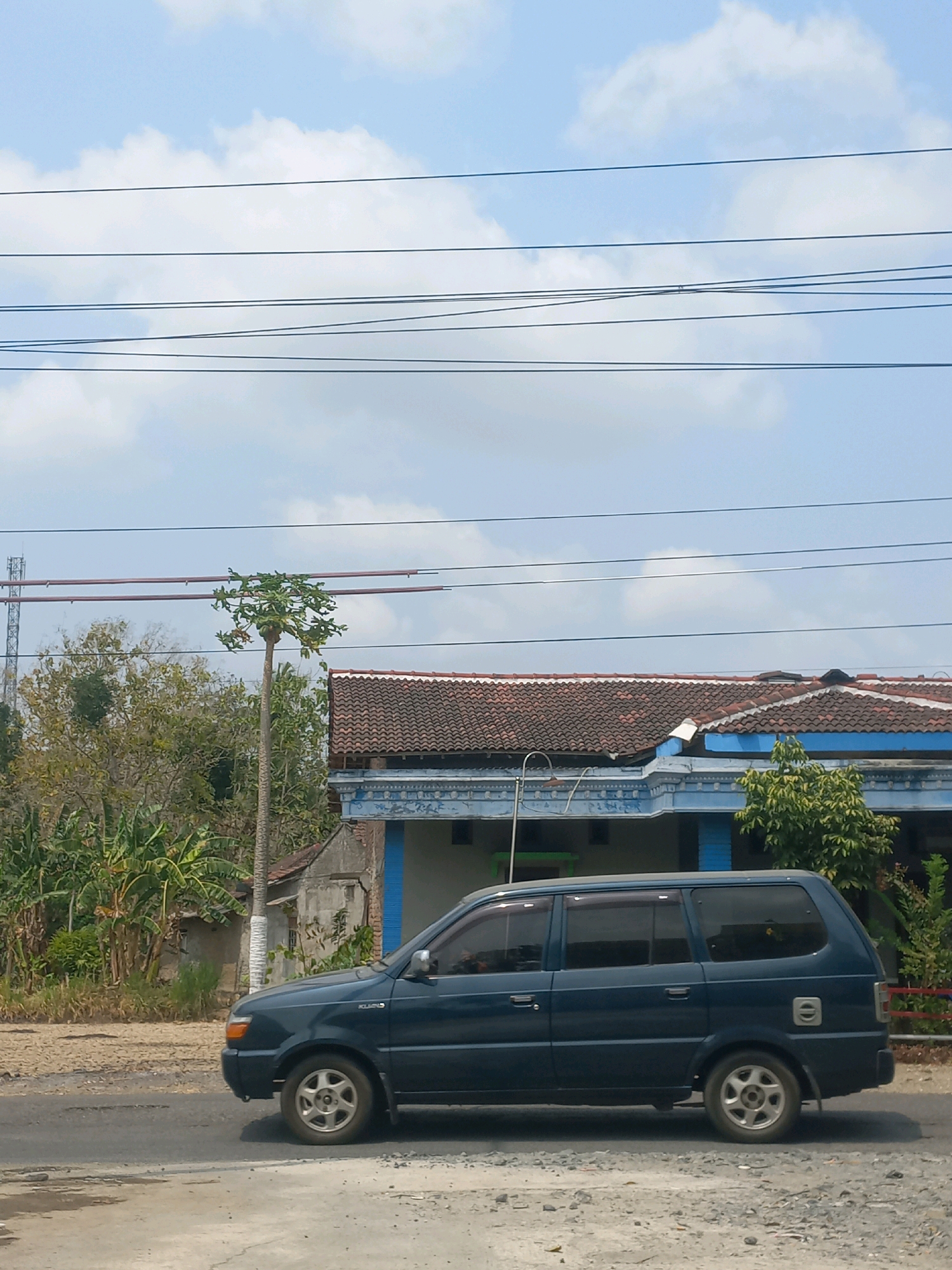 The image depicts a dark blue van driving on a paved road in Baturetno, Indonesia. The van is positioned in the right half of the frame, viewed from the side, with its front wheel visible and the rear wheel partially obscured. In the background, a single-story building with a tiled roof and blue trim is visible, suggesting a residential or commercial area. A tall, slender tree with a sparse canopy stands to the left of the building, and dense foliage with banana trees is present behind it. Overhead, power lines traverse the sky, and a telecommunications tower is visible in the far distance on the left. The sky is partly cloudy, with a bright, sunny atmosphere. The road surface appears to be a mix of asphalt and gravel.