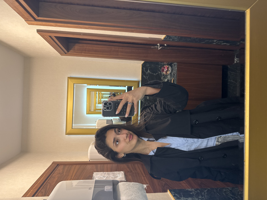 The image depicts a female subject taking a mirror selfie in an indoor setting. The specific location in Budva, Montenegro, is not discernible from the visual evidence.
The subject is a woman with long, dark hair, wearing a light blue striped collared shirt under a black trench coat. She has small hoop earrings and a ring on her left ring finger. She holds a dark-colored smartphone with a triple-lens camera array in her left hand, positioned to capture her reflection. Her lips are slightly pouted.
The immediate environment visible in the reflection includes a light-colored wall with dark reddish-brown wooden paneling or doors on both the left and right sides. On the left, a white, wall-mounted paper towel dispenser with a roll of white paper towels is visible. Below the subject's reflection, a dark green or black marble-patterned countertop with a dark wooden cabinet underneath is present. On the countertop, to the right of the subject, a clear glass container holding red, rose-like decorative elements is visible.
Behind the subject's reflection, a smaller mirror with a gold-colored frame is present, reflecting a similar mirror further in the background, creating a series of receding reflections. The bottom edge of the primary mirror in which the selfie is taken also features a gold-colored frame.