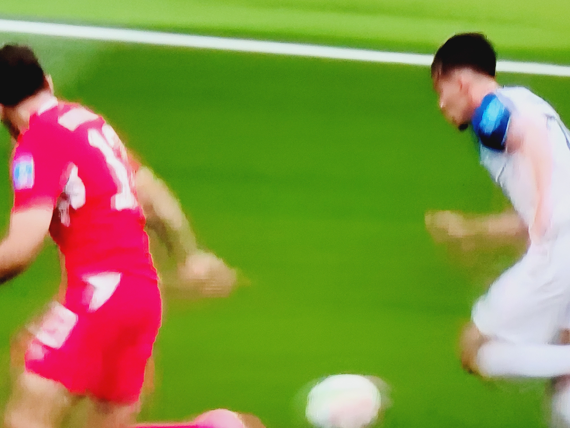 In Baku City, Azerbaijan, two male soccer players are captured in a dynamic action shot on a vibrant green soccer field. The player on the left, wearing a red jersey with white accents and the number "1" clearly visible, appears to be in motion, potentially kicking or dribbling a soccer ball. His body is turned away from the camera, and his face is not fully visible. To the right, another male player in a white jersey with blue accents is also in motion, facing towards the left. His pose suggests he is actively engaged in the game, possibly defending or attempting to intercept. A white and pink soccer ball is visible on the field between the two players. The background consists of the green field and a blurred white line, indicating the boundary of the playing area. The image captures a moment of intense gameplay, with the players' movements slightly blurred due to the action.
