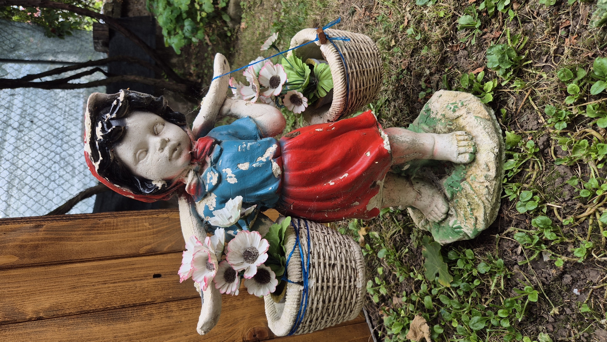 A weathered garden statue of a young girl carrying baskets of artificial flowers on her shoulders stands on a patch of green ground. The statue is made of concrete and is painted with red, blue, and white colors. The girl wears a red hood, a blue shirt, and a red skirt. Her face is tilted down, and her eyes are closed. The baskets she carries are woven from natural materials and contain white artificial flowers with pink tips and green leaves. The statue is placed in a garden setting, with a wooden fence to the left and trees and foliage in the background. The ground is covered in grass and soil, with some fallen leaves. The overall scene suggests a rustic and somewhat neglected outdoor space, possibly in a residential area. The image is taken at a medium distance, with the statue being the main focal point. No specific location in Doirentsi, Bulgaria is identifiable from the image itself.